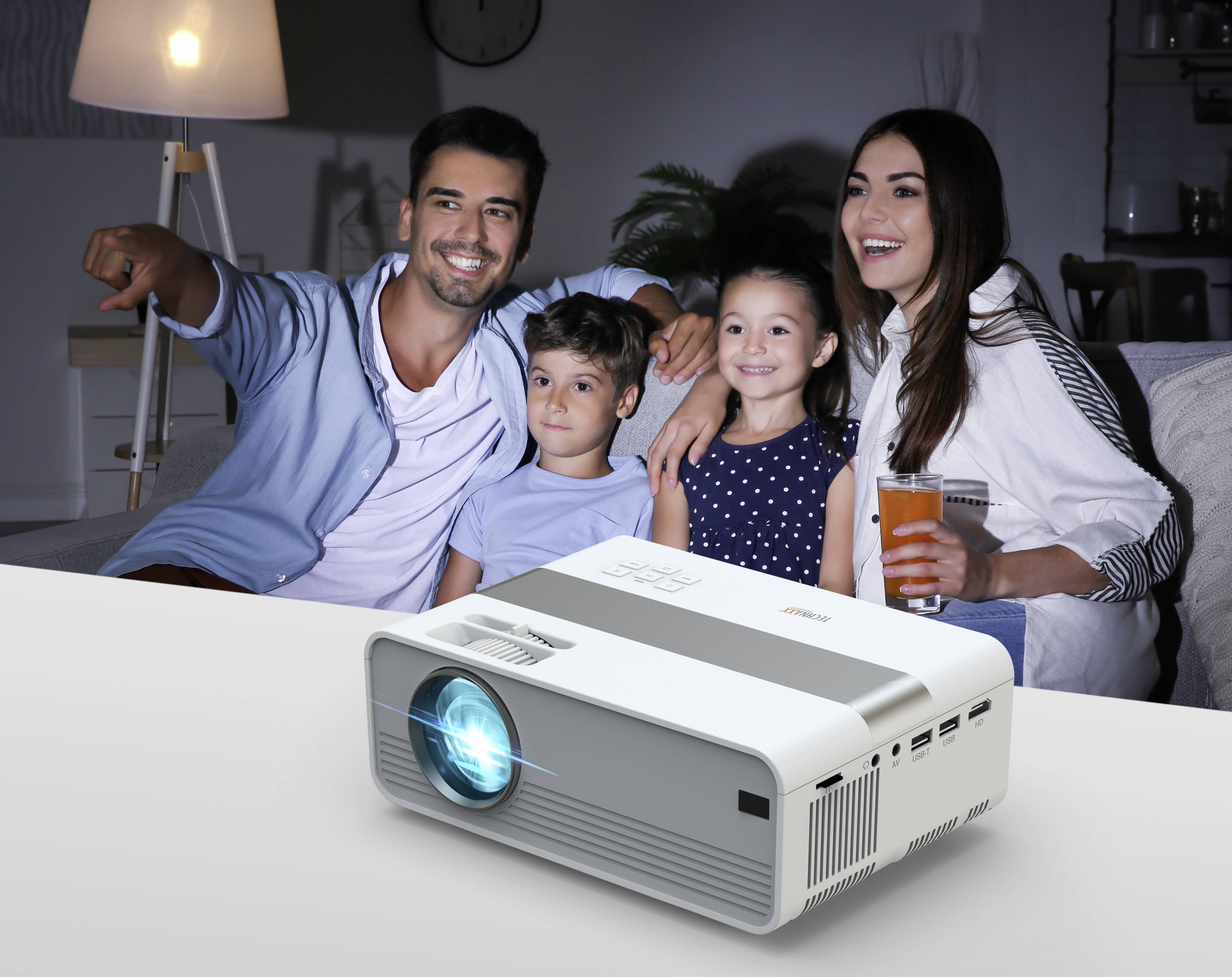 Eine Familie mit zwei Kindern sitzt lächelnd auf einem Sofa und schaut einen Film. Ein Projektor auf dem Tisch strahlt ein blaues Licht aus.