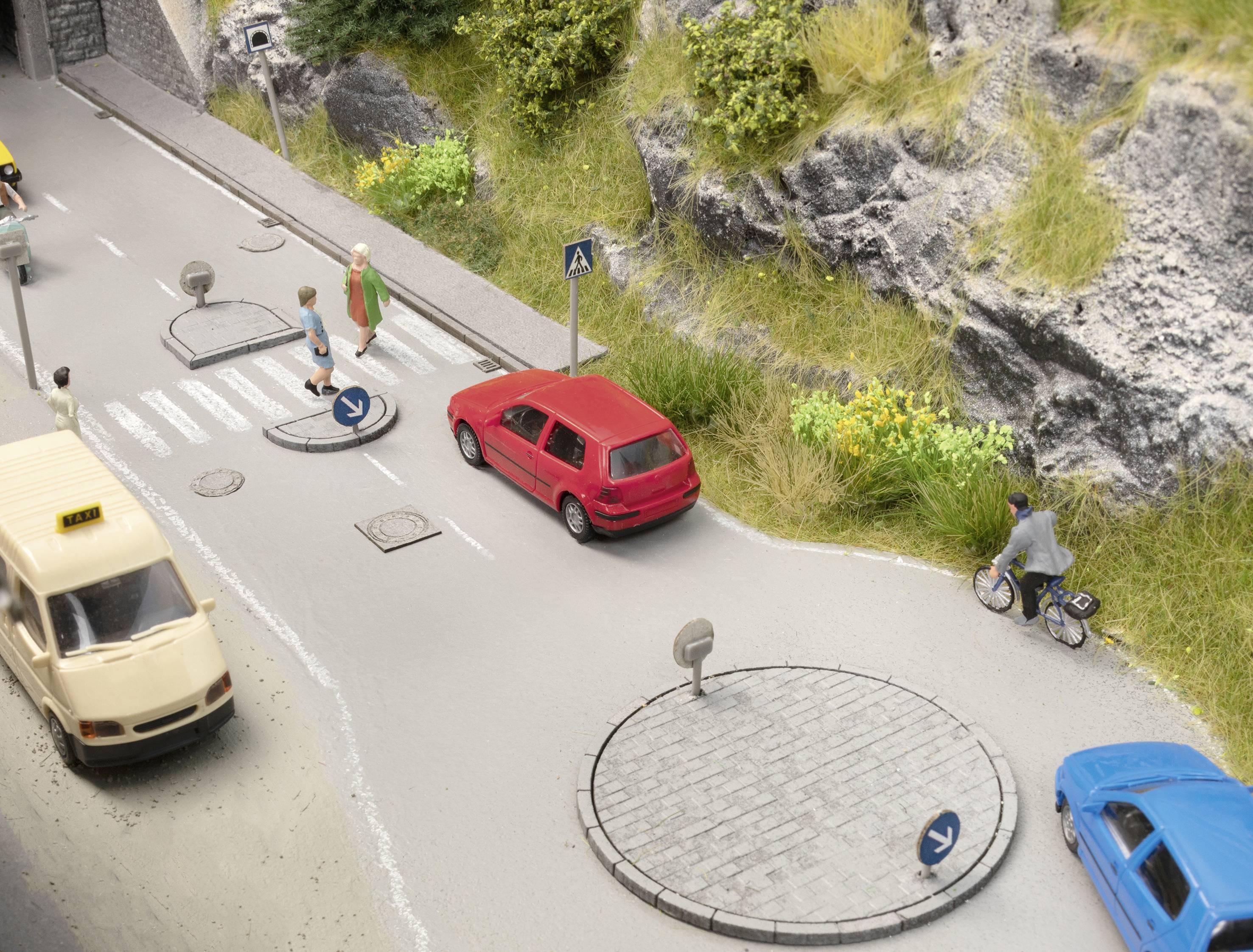 Modellstraße mit Miniaturautos, Fußgängerüberweg, rotem Auto und Radfahrer auf Fahrradweg entlang einer felsigen Böschung.
