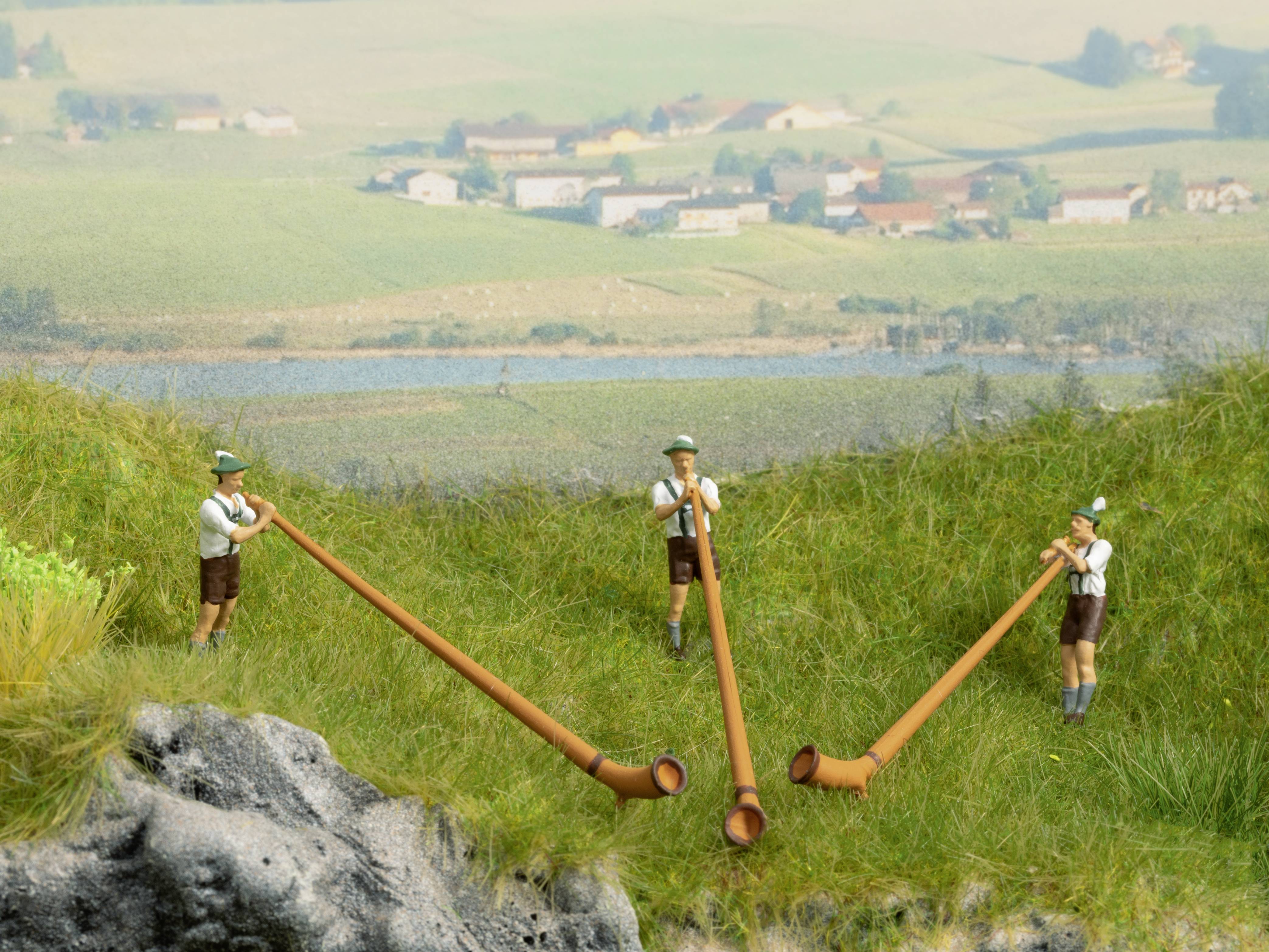 Drei Figuren in Tracht spielen Alphörner auf einer grünen Wiese mit einem Dorf und Feldern im Hintergrund.