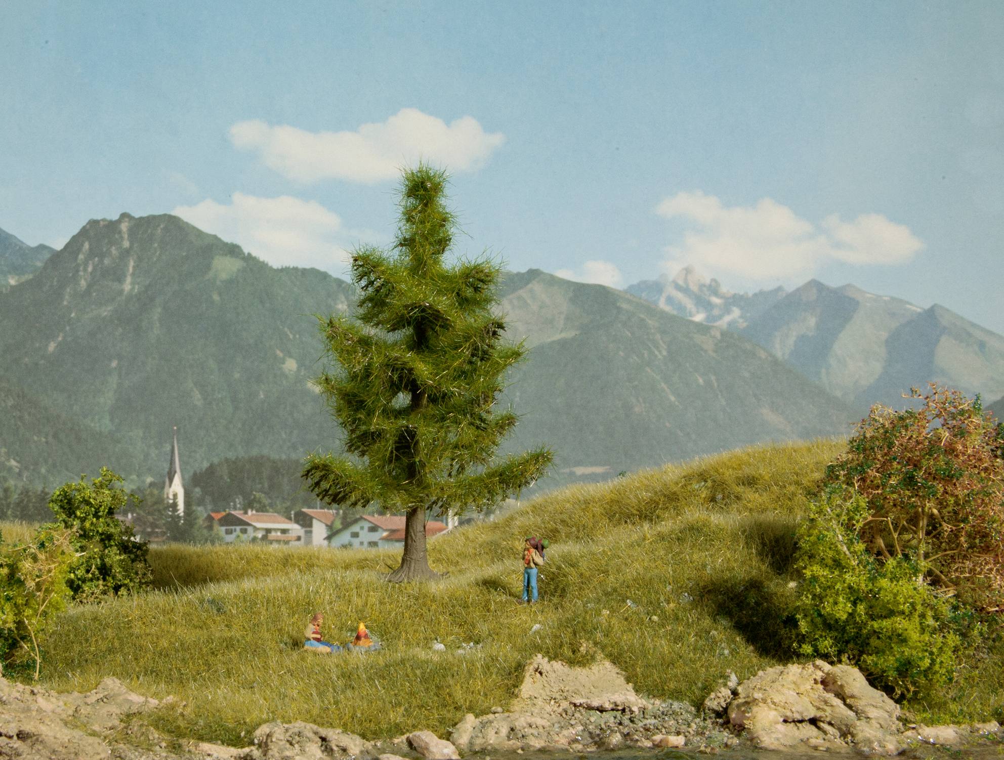 Eine Miniaturlandschaft zeigt ein Dorf in den Bergen. Zwei Figuren sitzen auf einer Wiese vor einem Baum, im Hintergrund Berggipfel und ein Kirchturm.