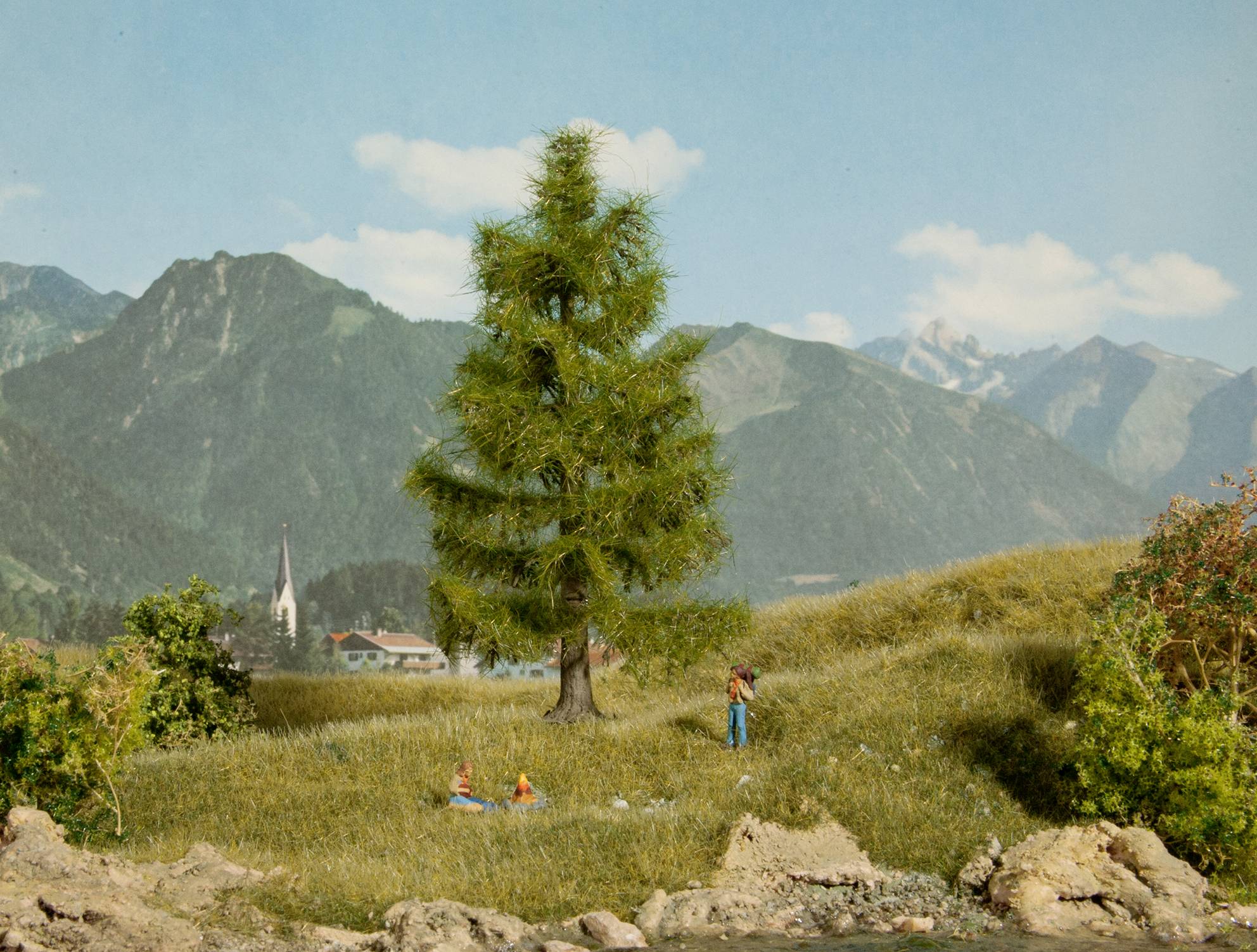 Ein großes Modell eines einsamen Baumes in einer Graslandschaft. Im Hintergrund sind Miniaturmenschen und eine Miniaturkirche zu sehen.