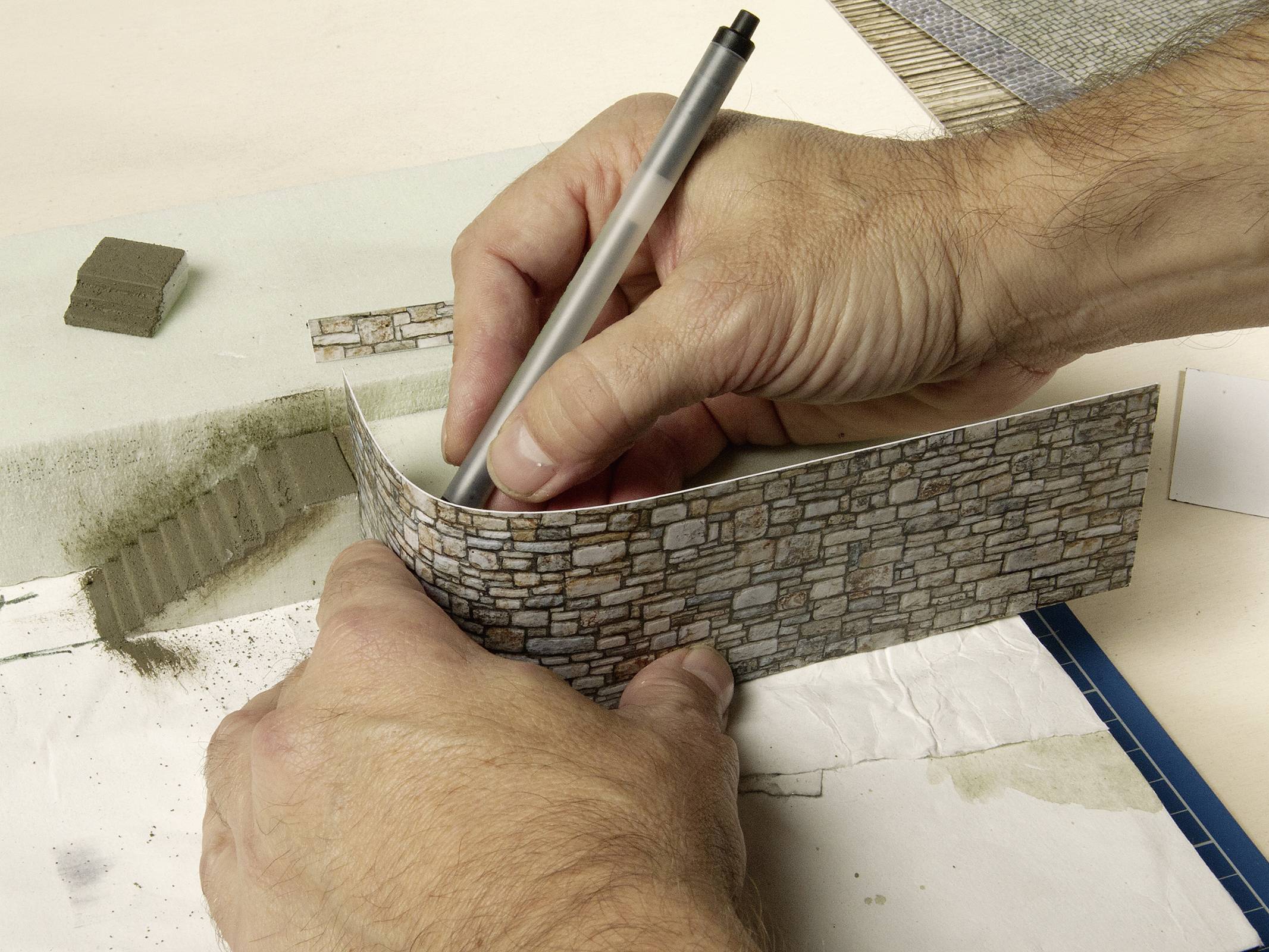 Zwei Hände bauen ein Miniatur-Diorama, befestigen gedruckte Steinmauer an der Form mit einem Stift und Klebstoff.