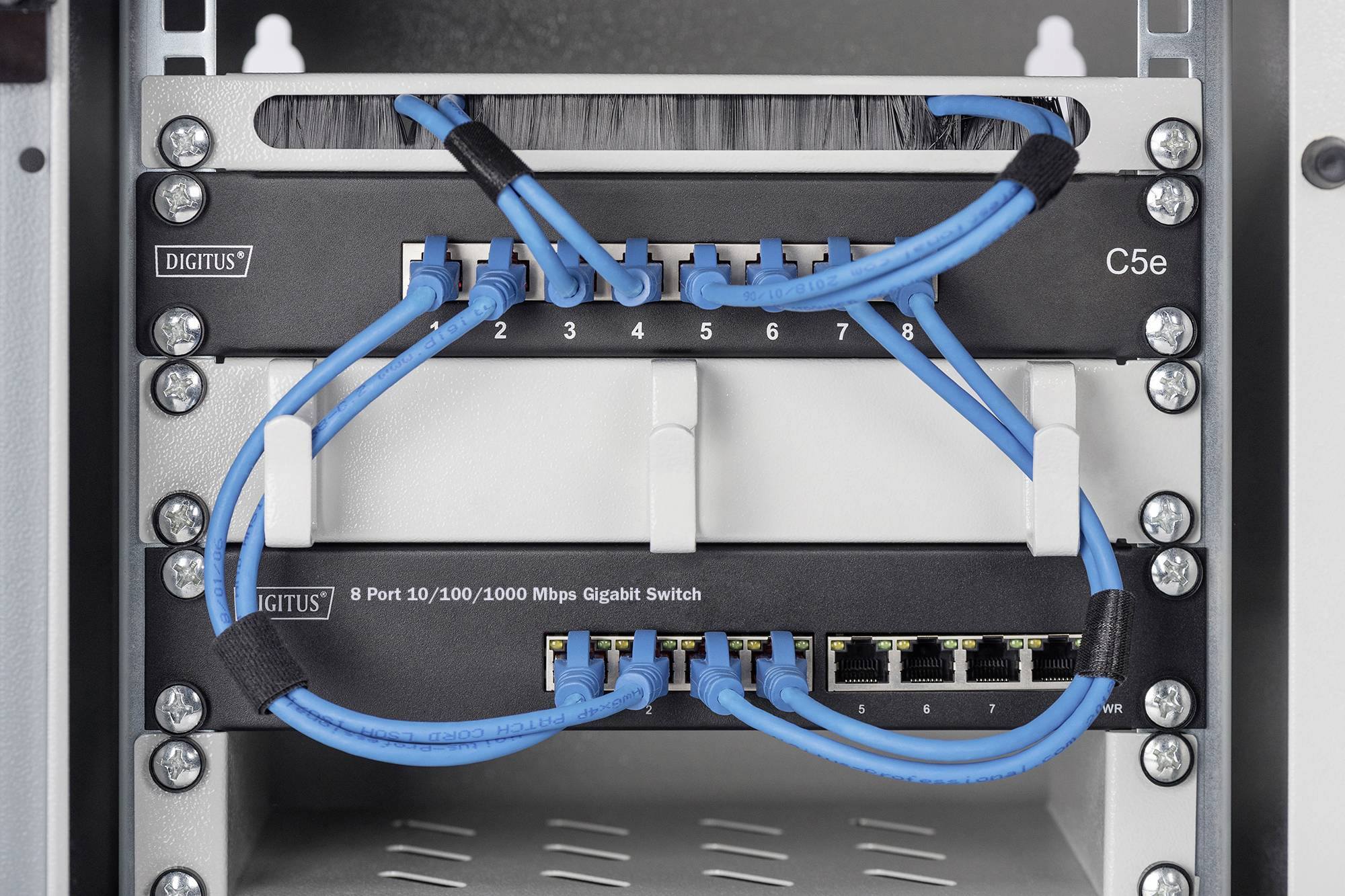 Netzwerk-Switch im Schrank zeigt acht angeschlossene blaue Ethernet-Kabel. Beschriftung lautet '8 Port 10/100/1000 Mbps Gigabit Switch'.
