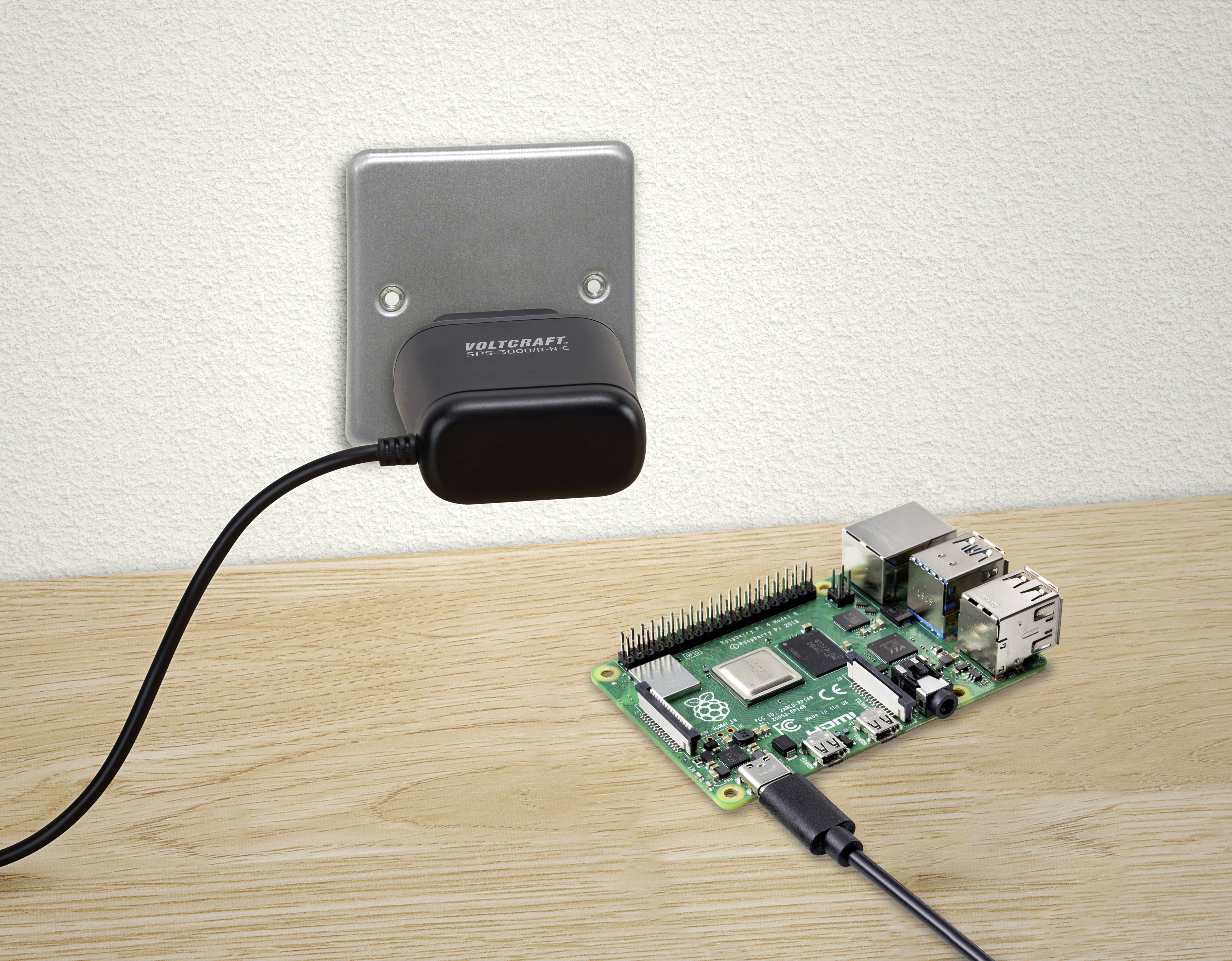 Ein Raspberry Pi ist auf einem Holztisch zu sehen, verbunden mit einem Netzteil, das in einer Steckdose steckt.