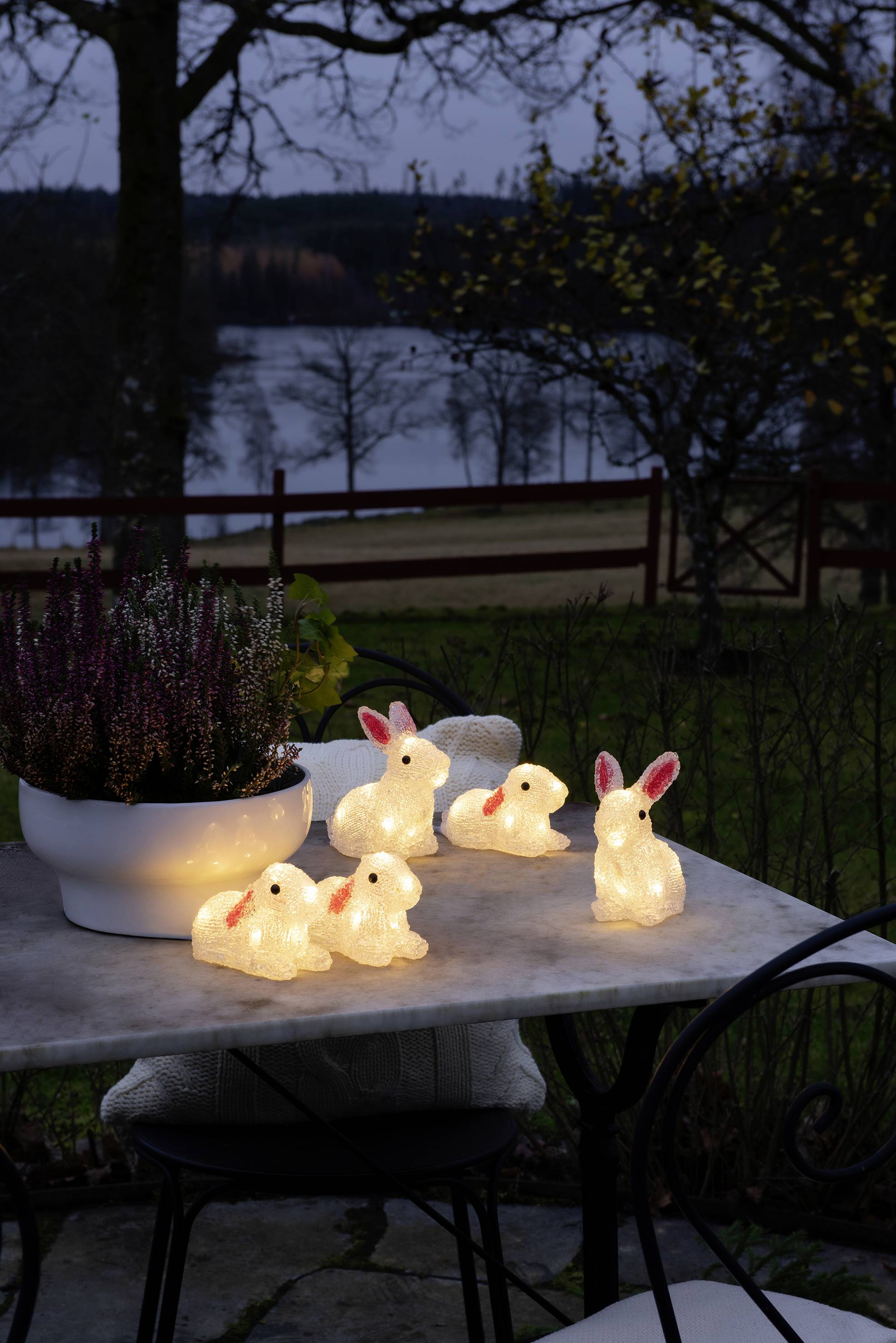Leuchtende Hasenfiguren auf einem Tisch bei Dämmerung, umgeben von Pflanzen. Im Hintergrund ein See und ein Baum. Stimmungsvolle Gartendekoration.