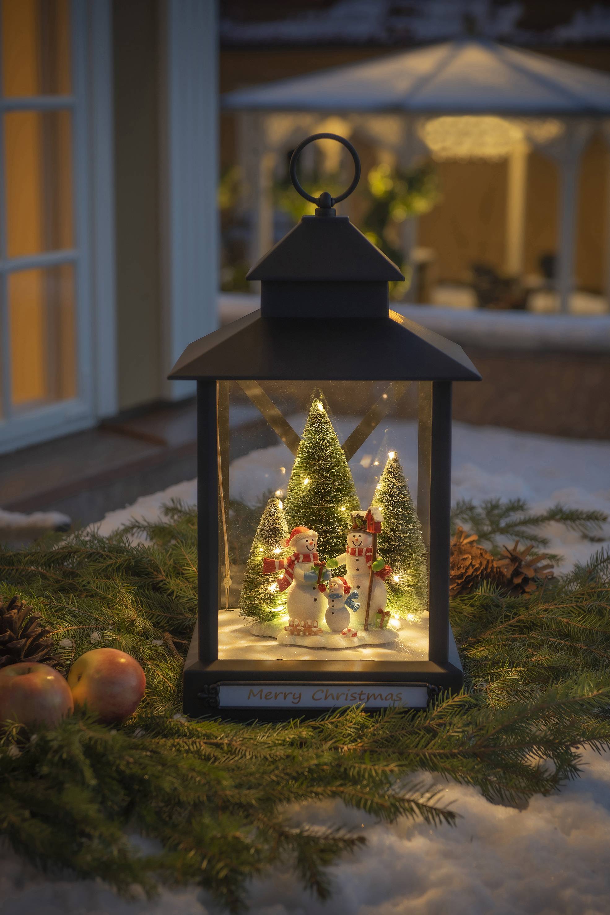Eine Laterne mit Weihnachtsszene: beleuchtete Schneemänner und Tannenbäume stehen auf Kunstschnee, umgeben von Äpfeln und Tannenzweigen.