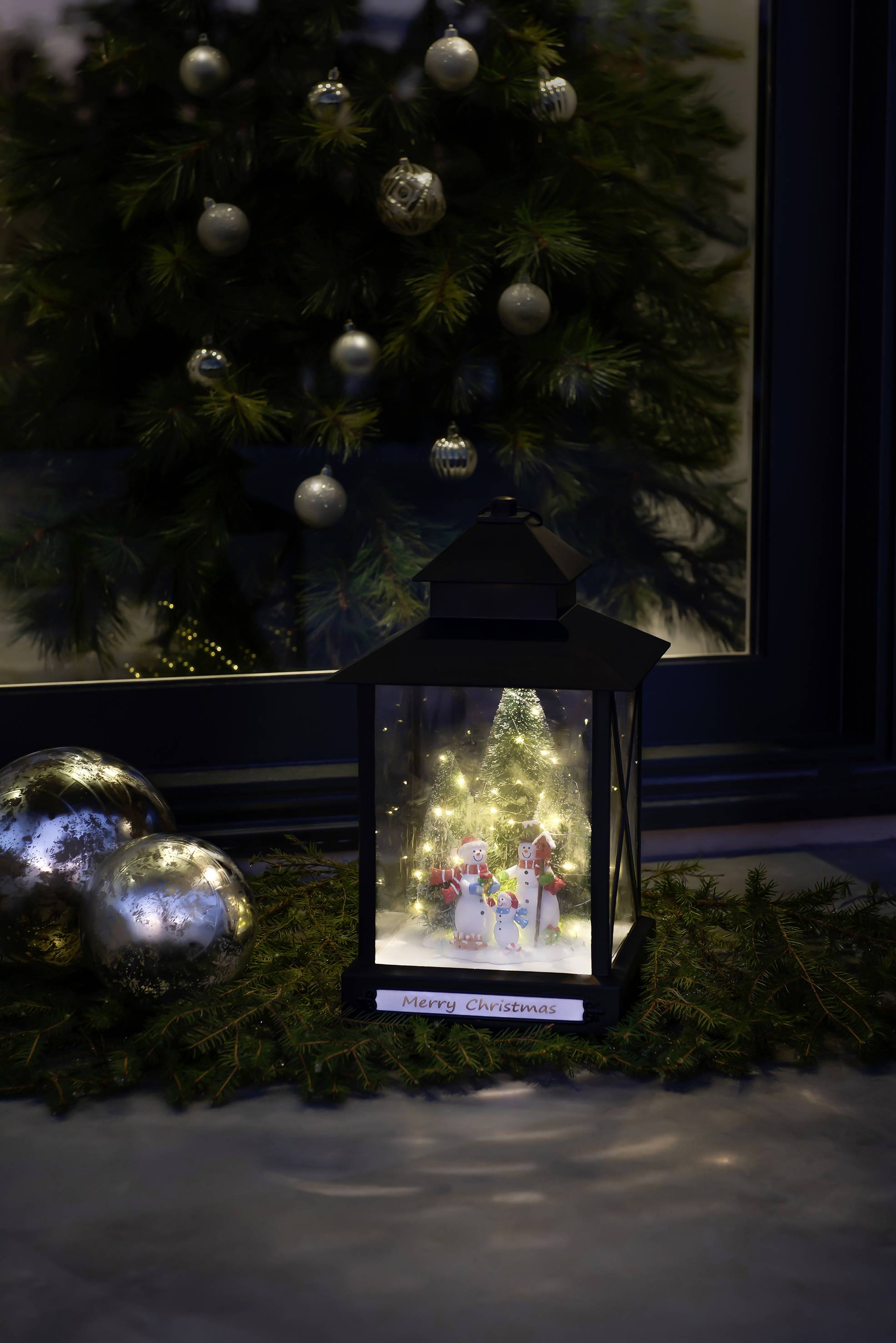 Laterne mit Weihnachtsdekoration, beleuchtet vor einem Fenster mit Weihnachtsbaum und Kugeln. Text auf der Laterne: 'Merry Christmas'.
