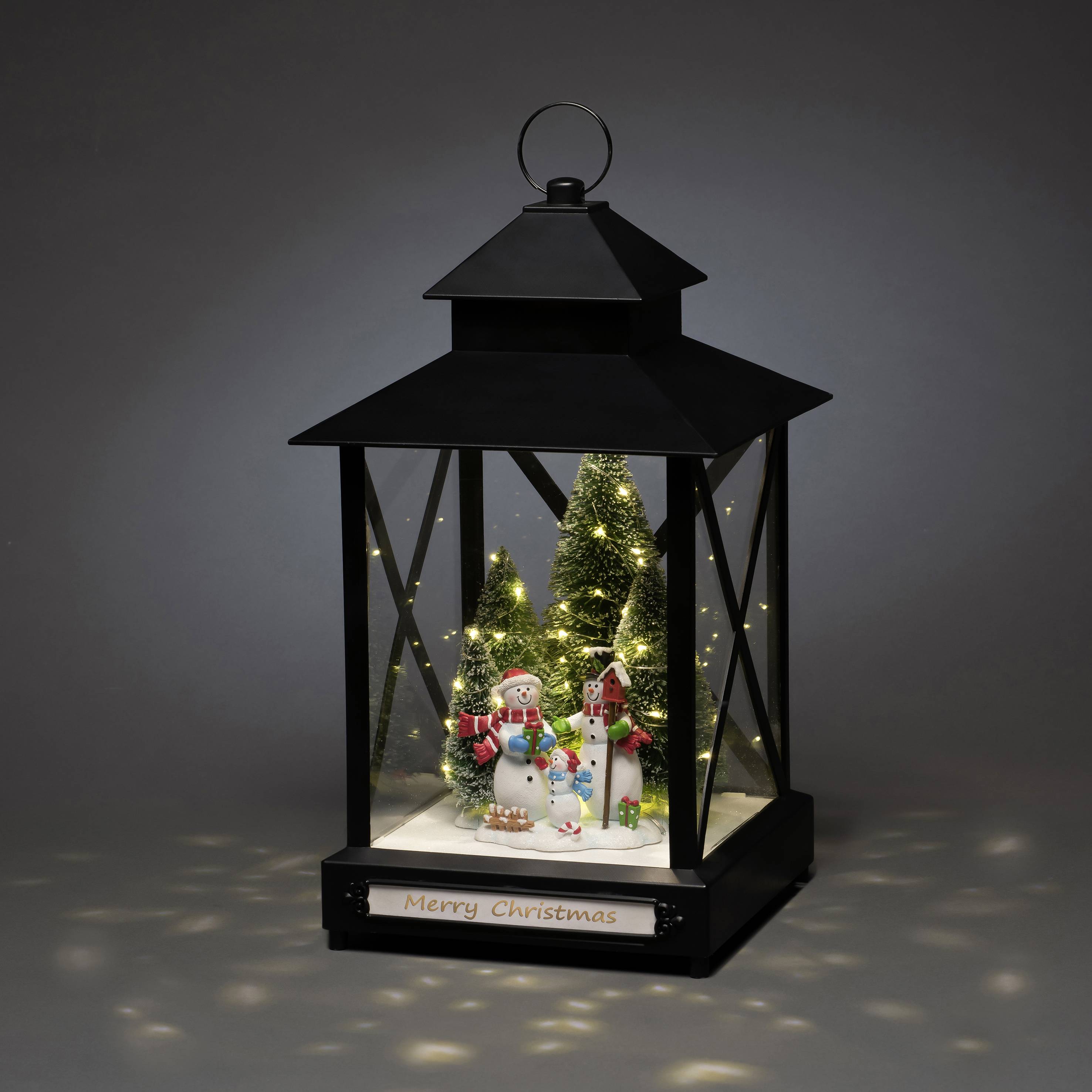 Eine schwarze Laterne mit Schneemann-Deko und Weihnachtsbäumen im Inneren unter einer LED-Beleuchtung. Aufschrift 'Merry Christmas'.