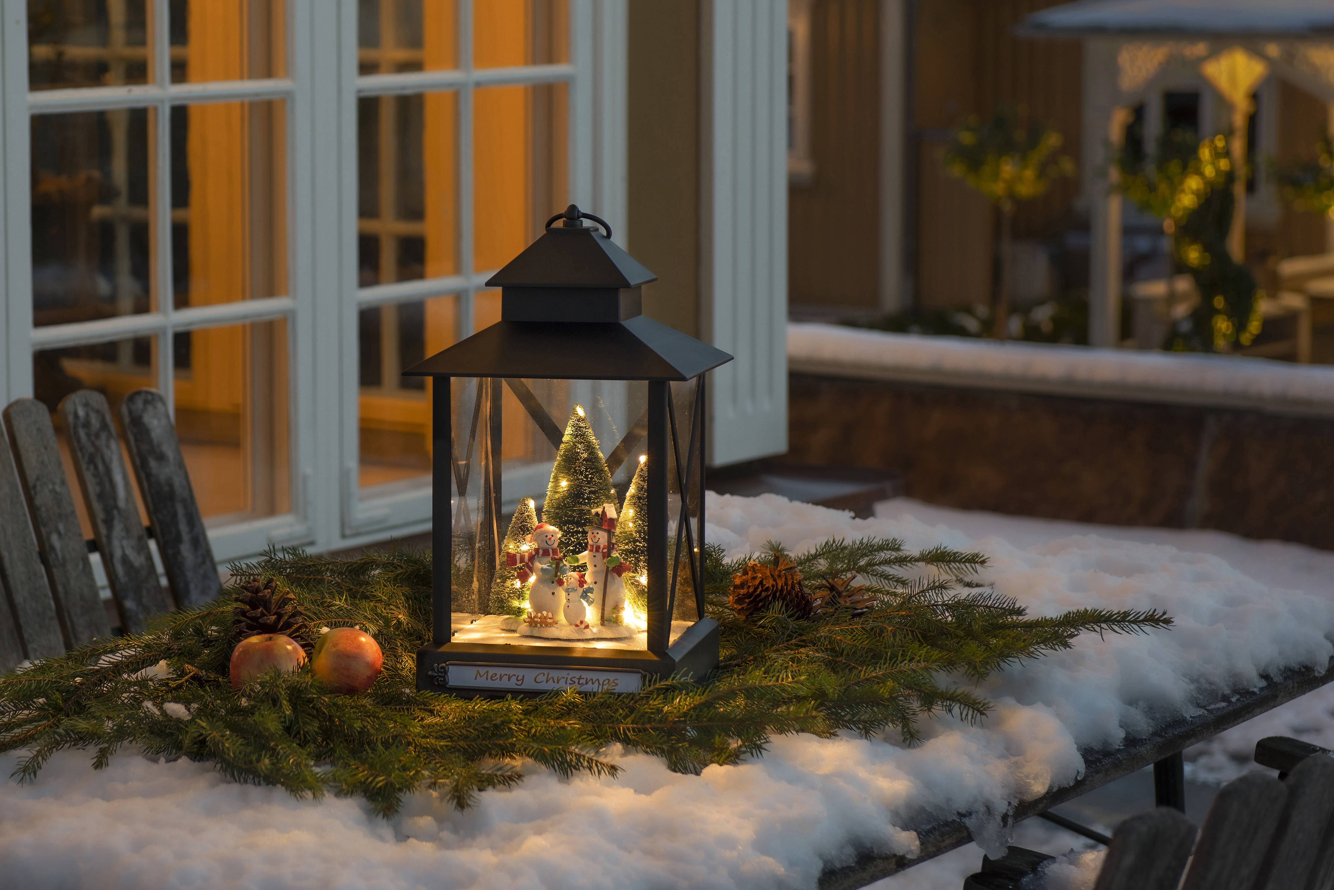 Eine beleuchtete Laterne mit Weihnachtsdekoration steht auf einem schneebedeckten Tisch, umgeben von Kiefernzweigen und Ornamenten.