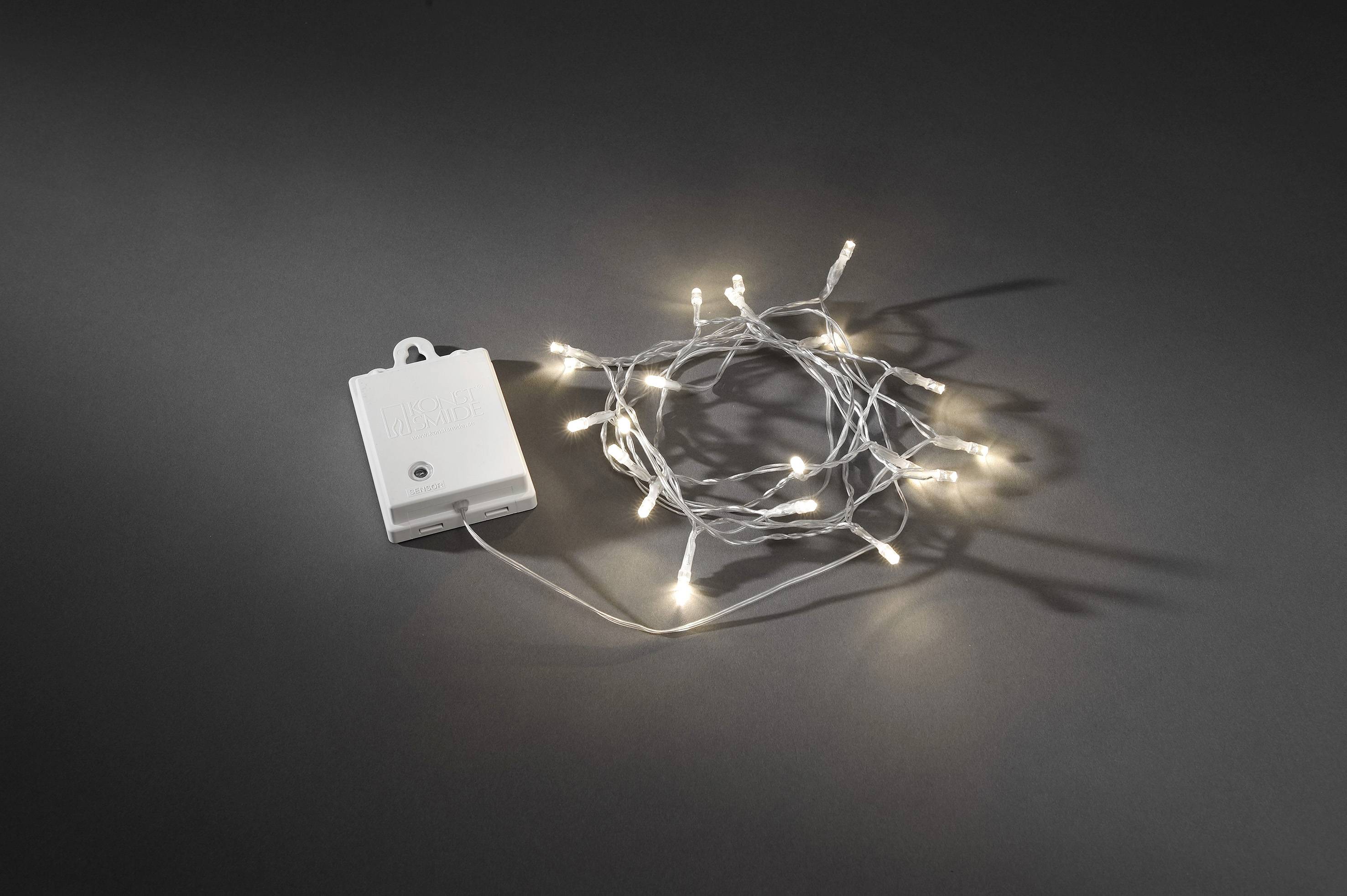 Lichterkette mit kleinen, leuchtenden LED-Leuchten, aufgerollt neben einem rechteckigen Batteriefach auf dunklem Hintergrund.