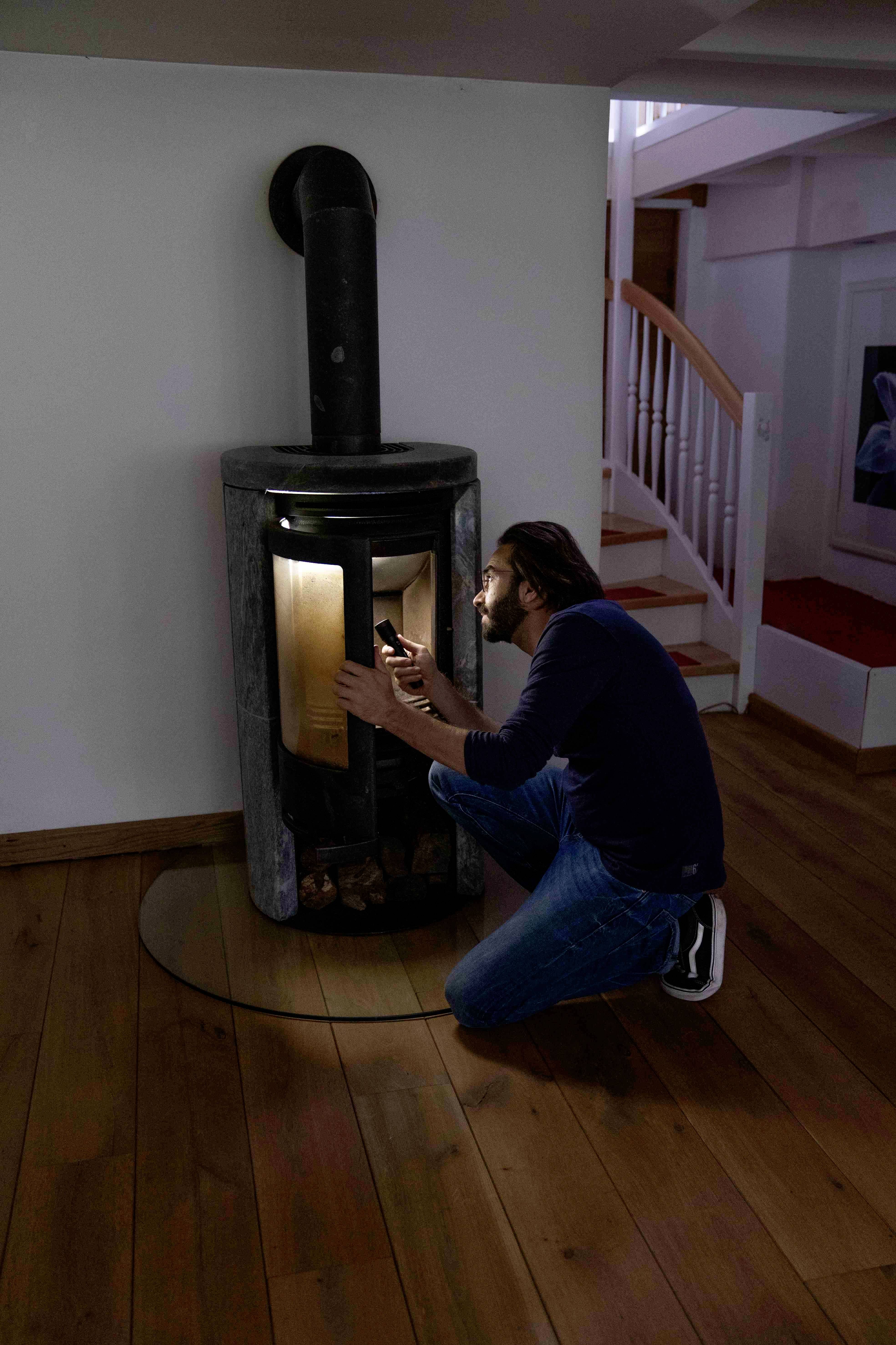 Eine Person sitzt auf dem Boden und entzündet ein Feuer in einem modernen Kamin in einem gemütlichen Raum mit Holzboden.