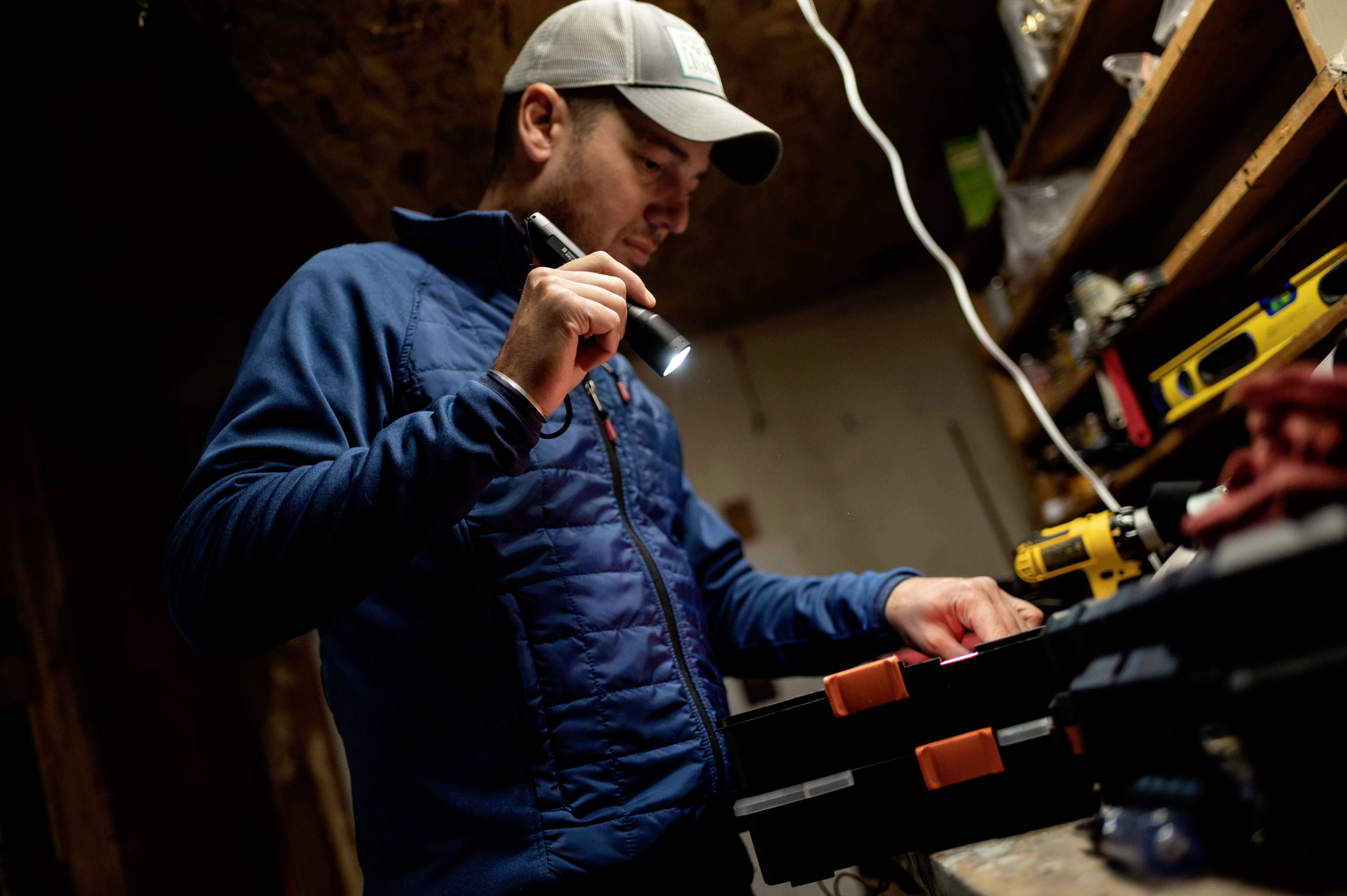 Ein Mann arbeitet in einer Werkstatt und hält eine Taschenlampe. Er trägt eine Mütze und eine blaue Jacke, umgeben von Werkzeugen.