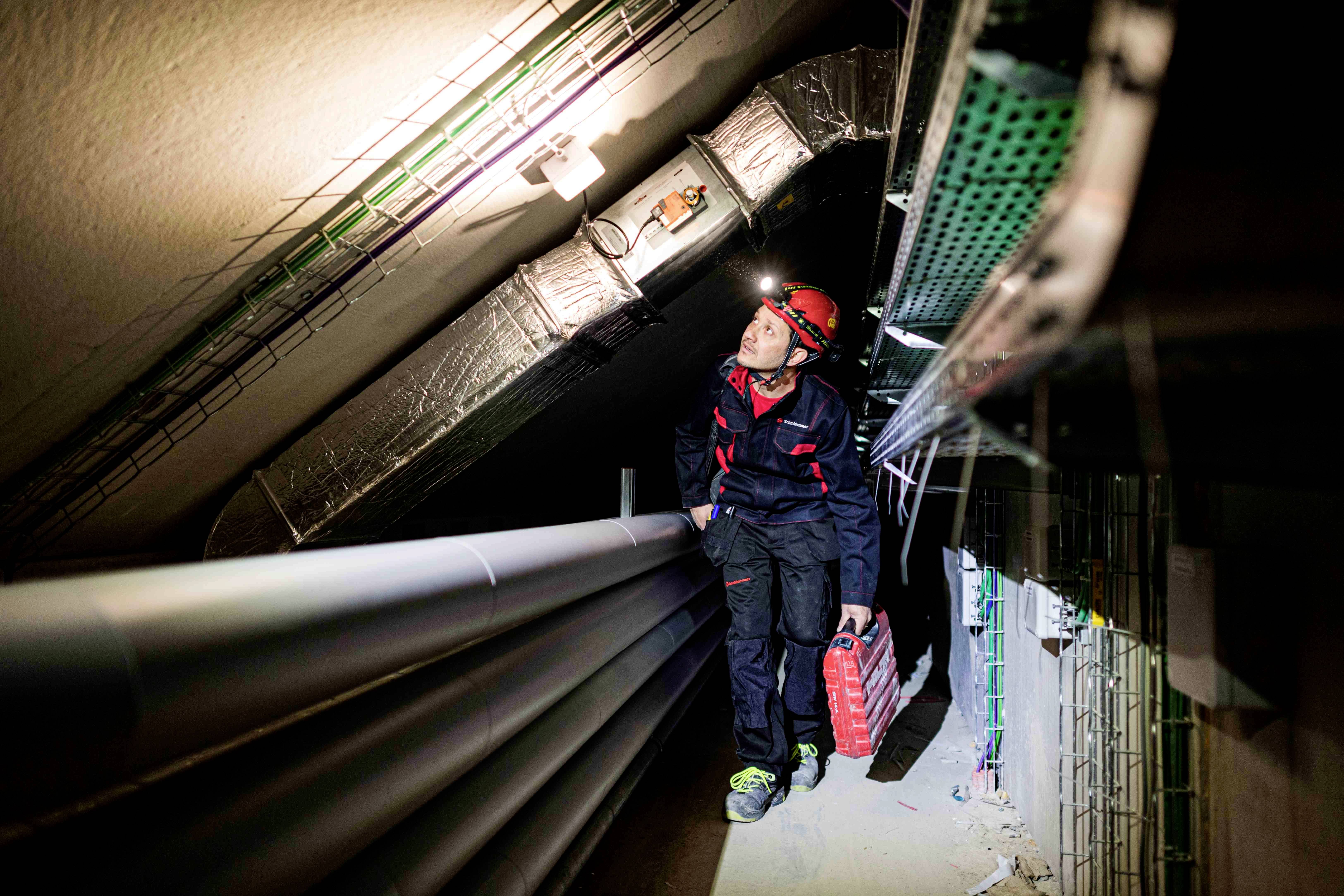 Ein Elektriker mit Helm und Werkzeugkoffer inspiziert Kabel in einem engen Tunnel, beleuchtet von einer Kopflampe.