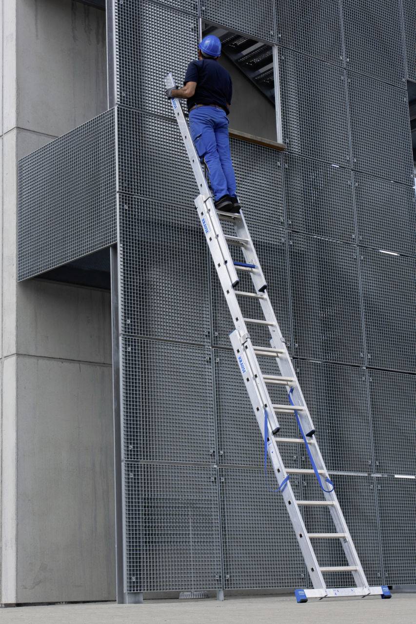 Ein Arbeiter in blauer Kleidung und Helm steht auf einer langen Leiter, die an einer Gebäude-Fassade angelehnt ist.