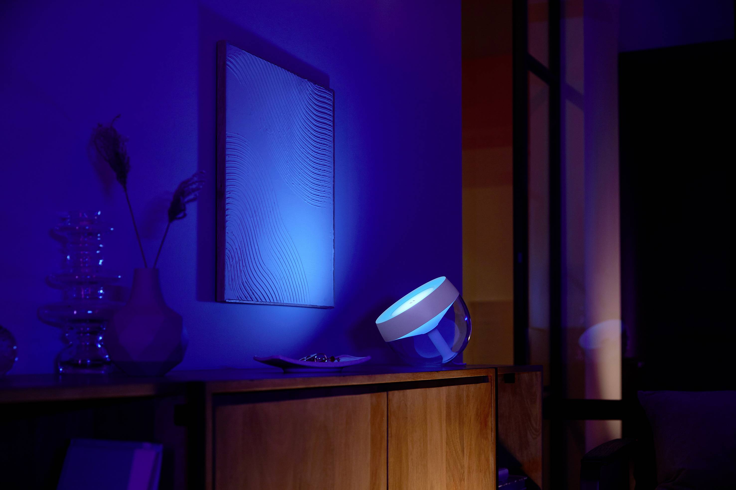 Ein Wohnzimmer mit dunkler Beleuchtung, ein blaues Licht strahlt auf ein Kunstwerk an der Wand. Der Raum wirkt modern und stimmungsvoll.