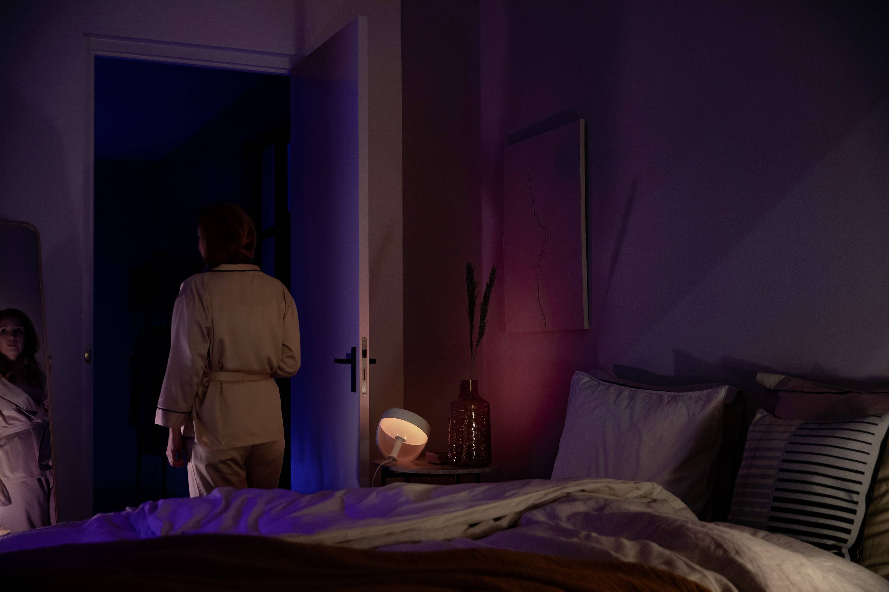 Eine Person im Pyjama steht in einem schummrig beleuchteten Schlafzimmer neben einem geöffneten Tür, das Bett ist ungemacht.