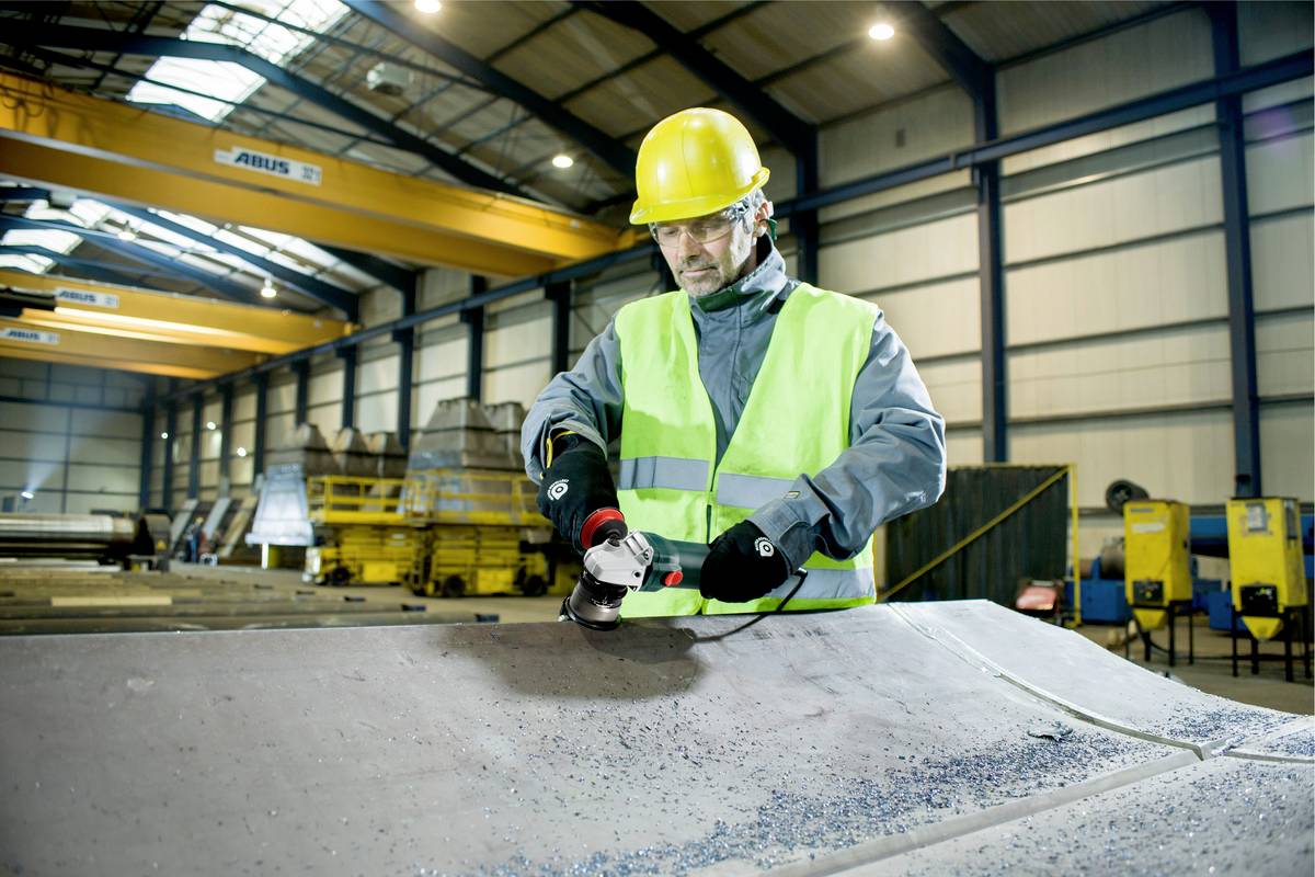 Ein Arbeiter in Sicherheitskleidung schleift Metall in einer großen Fabrikhalle. Im Hintergrund sind Kräne und Maschinen zu sehen.