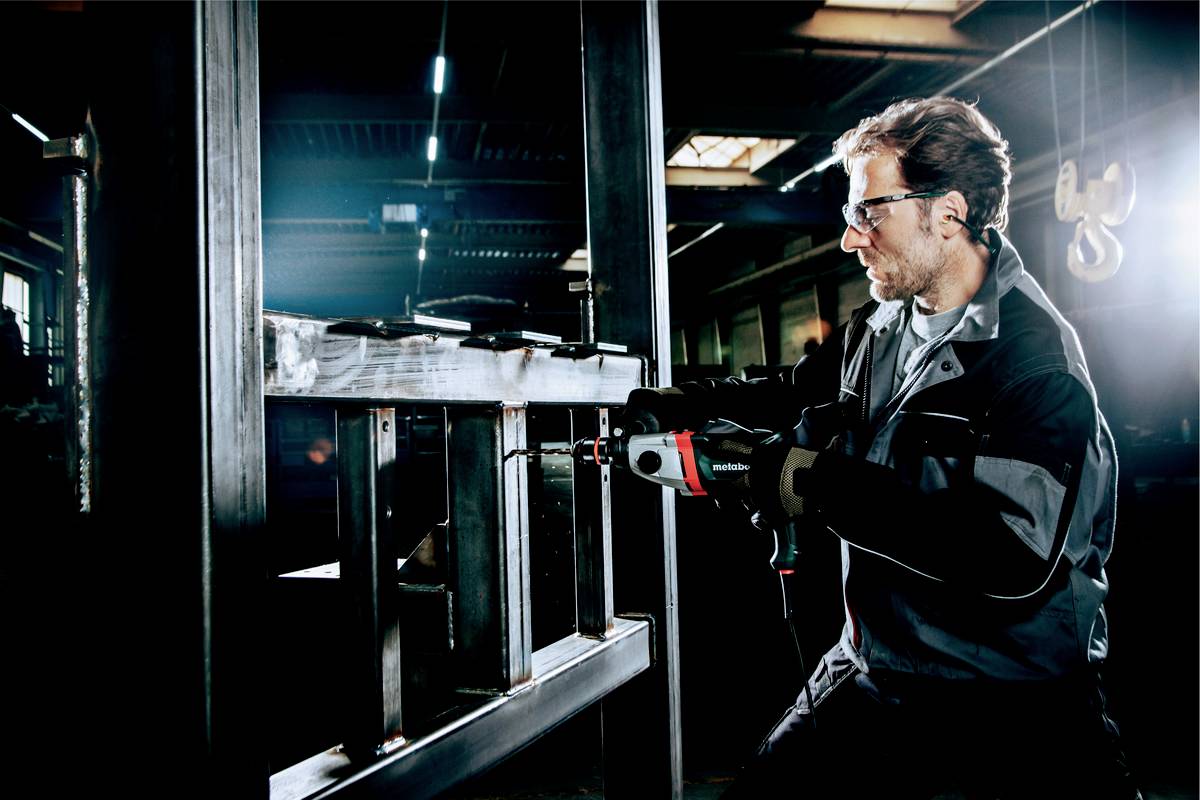 Ein Mann in Schutzkleidung arbeitet konzentriert mit einer Bohrmaschine an einem Metallgestell in einer industriellen Werkstattumgebung.