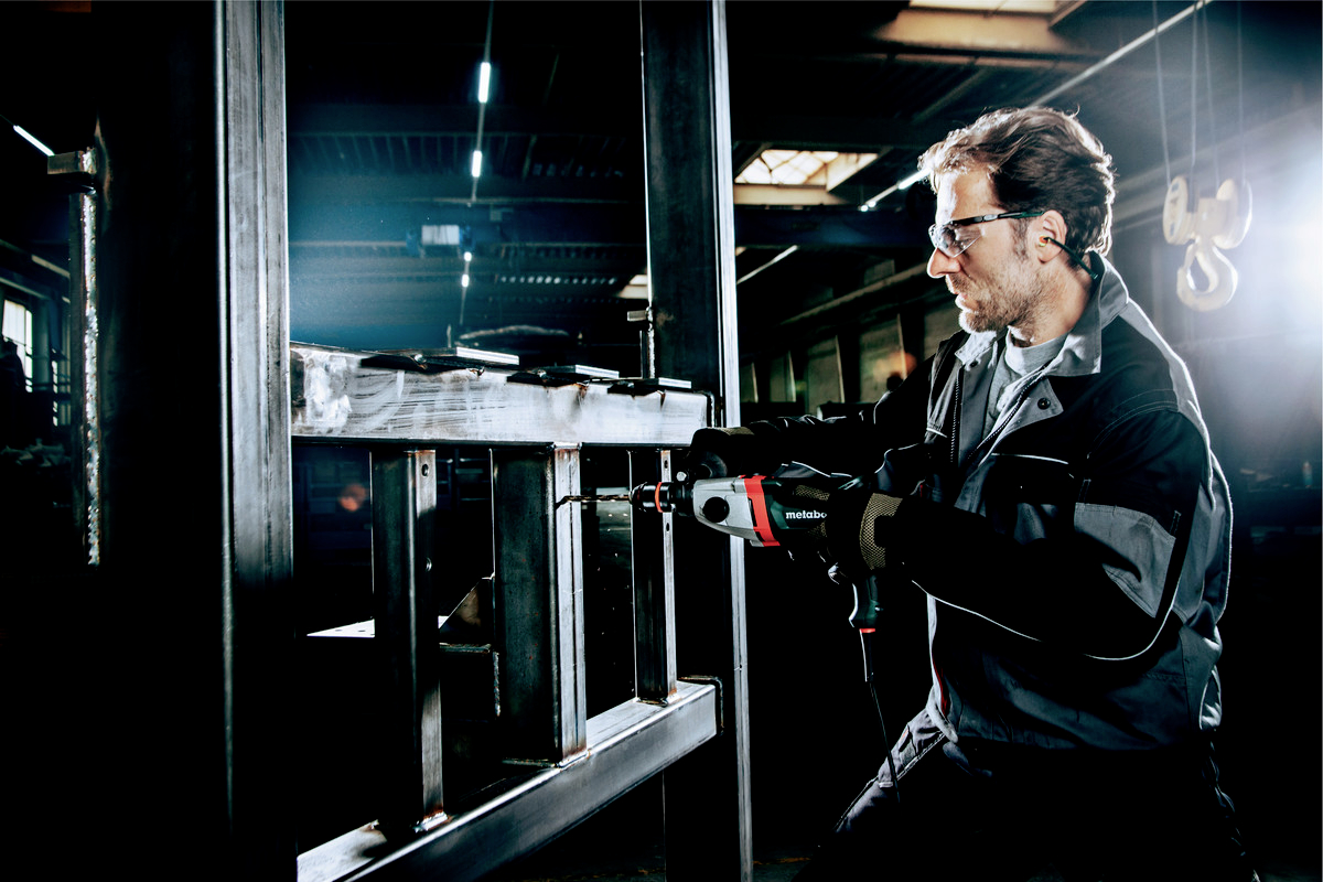 Ein Arbeiter mit Schutzbrille benutzt ein Elektrowerkzeug, um ein Metallgestell in einer Industriehalle zu bearbeiten.