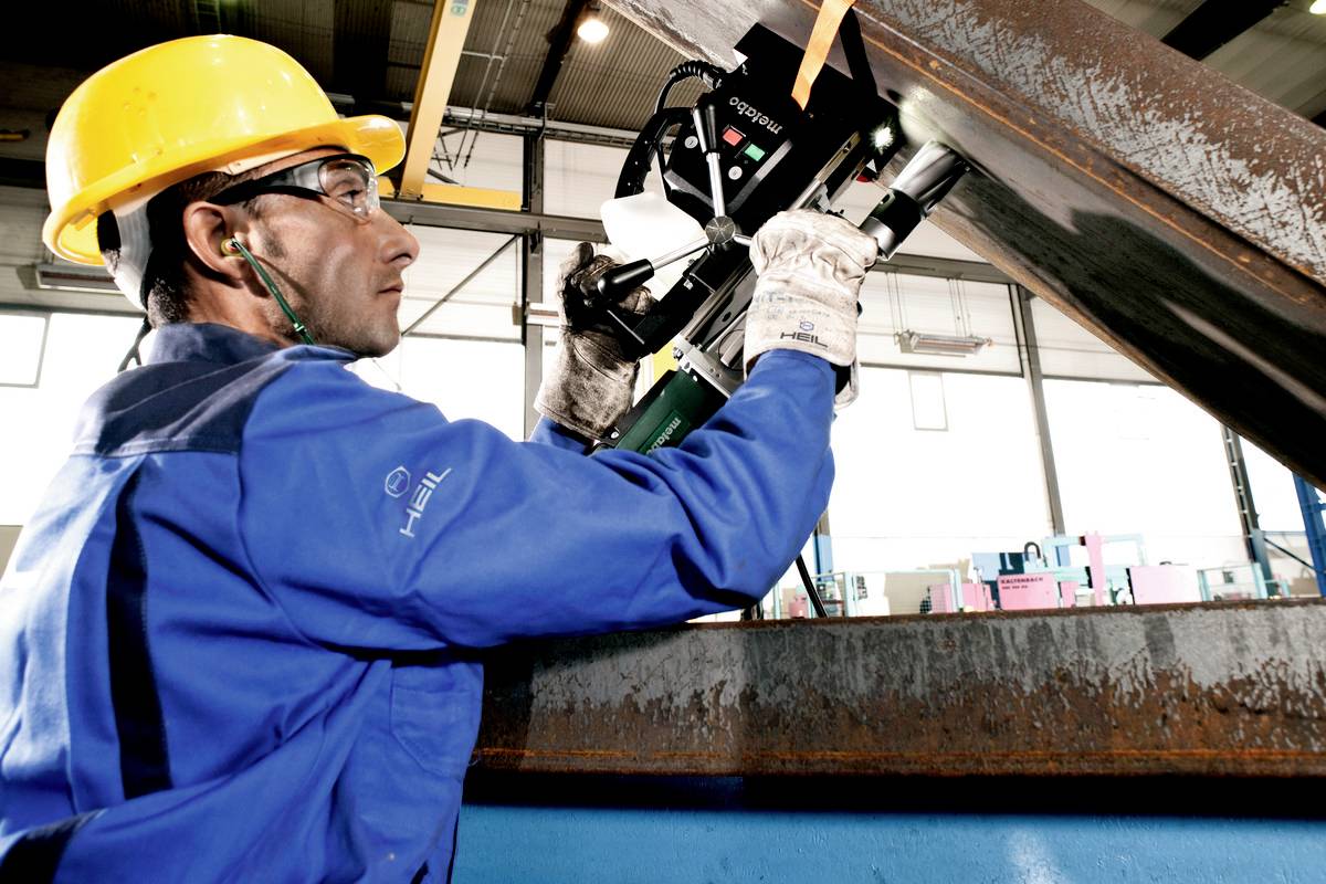 Ein Arbeiter in Schutzkleidung und Helm bedient ein handgeführtes Schneidgerät in einer Fabrikhalle. Die Umgebung ist industriell.