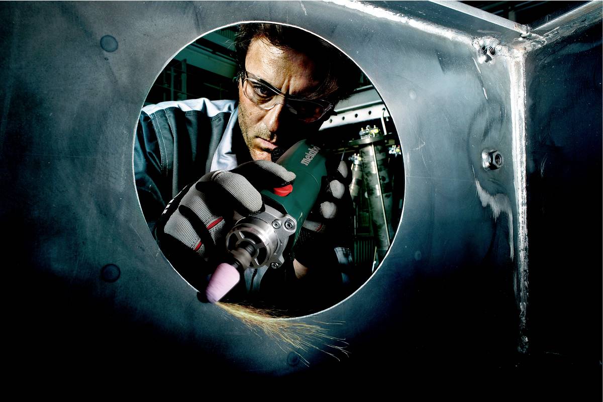 Ein Mann in Arbeitskleidung und Schutzbrille schleift mit einem Elektrowerkzeug Metall in einer industriellen Umgebung.