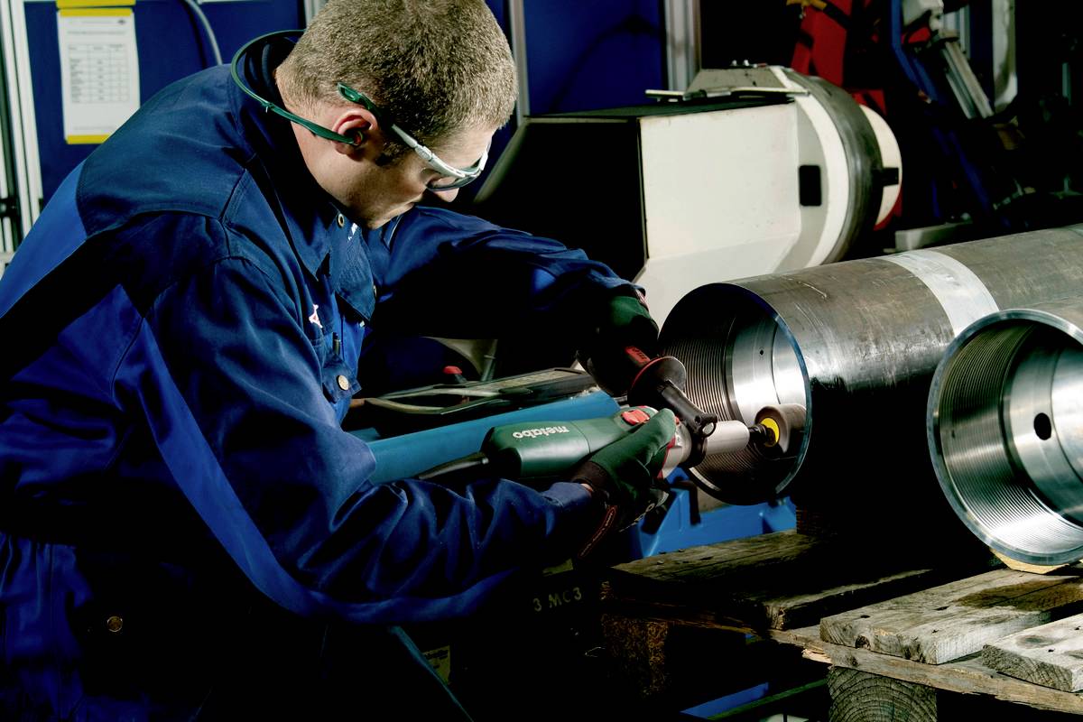 Ein Arbeiter in Schutzkleidung schleift mit einem Elektrowerkzeug die Innenseite eines großen Metallrohrs in einer Werkstatt.
