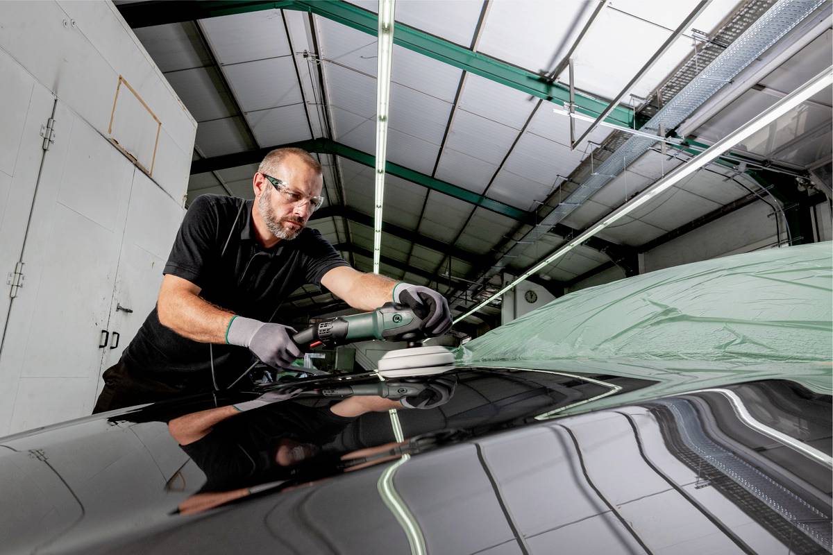 Ein Mann poliert das Dach eines Autos in einer Werkstatt mit einer Poliermaschine. Er trägt Schutzbrille und Handschuhe.