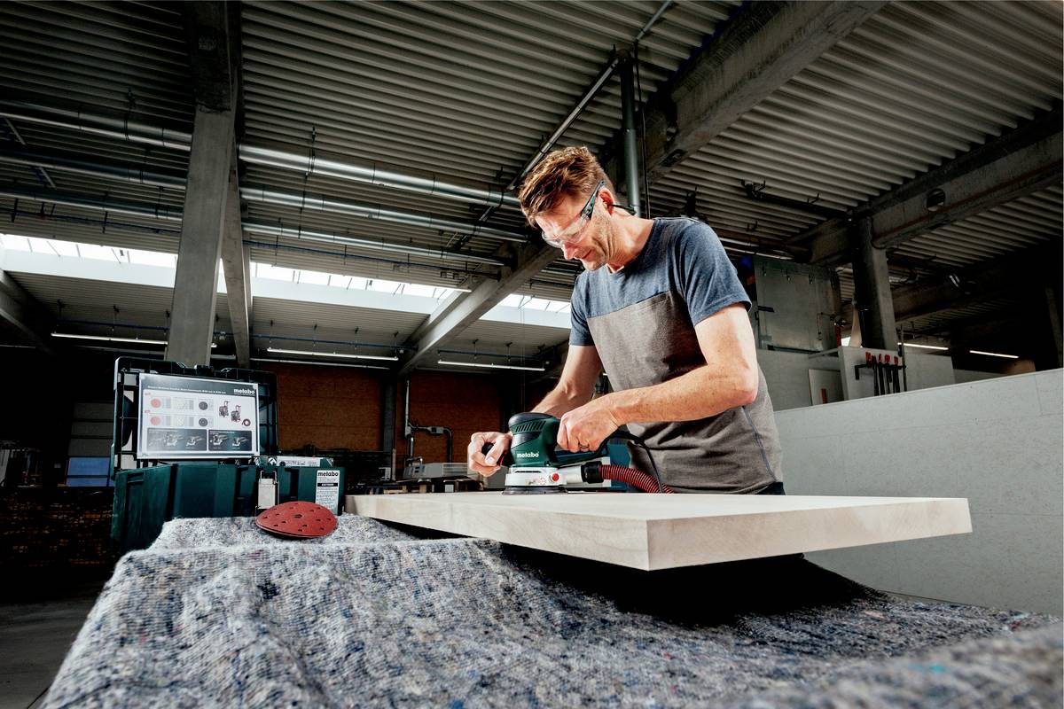Ein Mann schleift eine Holzplatte in einer Werkstatt. Er trägt Schutzbrille und Ohrstöpsel. Im Hintergrund ist Werkzeug sichtbar.