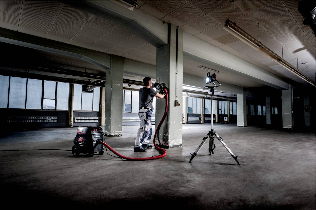 Eine Person arbeitet in einem großen, leeren Raum mit einem Industriestaubsauger und einer Lichtquelle auf einem Stativ.