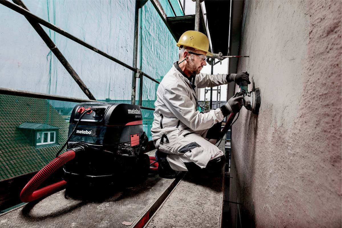 Ein Bauarbeiter mit Schutzhelm und Handschuhen schleift eine Wand auf einem Gerüst. Neben ihm steht ein Staubsauger.