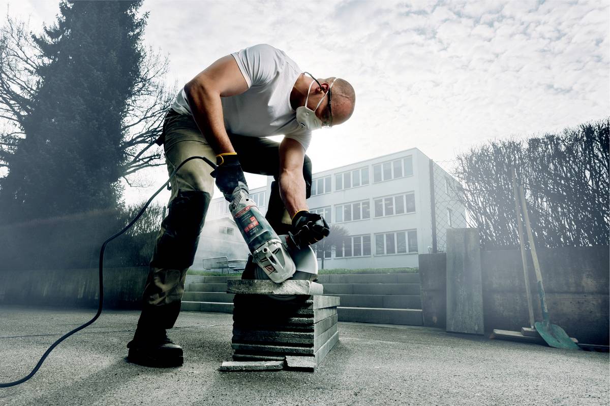 Eine Person mit Schutzbrille schneidet mit einem Winkelschleifer eine Steinplatte im Freien. Im Hintergrund sind Gebäude sichtbar.
