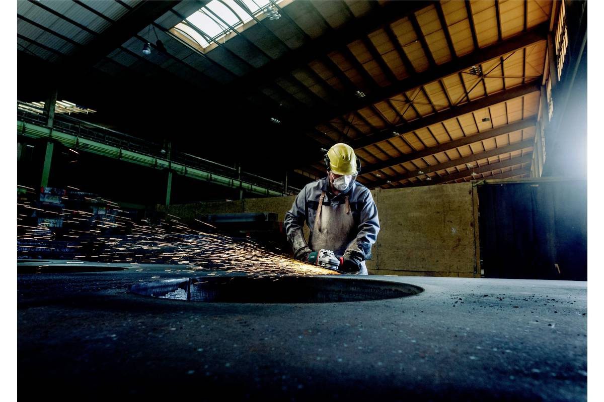 Ein Arbeiter in Schutzkleidung schleift Metall in einer Fabrikhalle, Funken sprühen. Hohe Decke und Lichteinfall von oben.