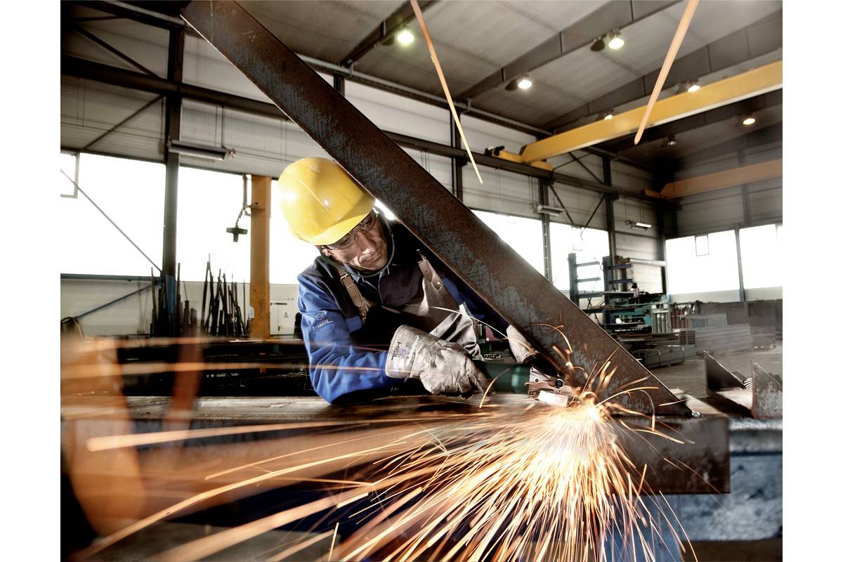 Ein Arbeiter in einer Werkhalle schleift Metall, wodurch Funken entstehen. Er trägt Schutzkleidung und einen gelben Helm.