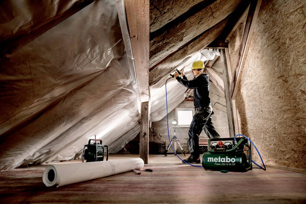 Eine Person mit gelbem Schutzhelm arbeitet mit einem Elektrowerkzeug in einem ausgebauten Dachboden. Isoliermaterial ist sichtbar.