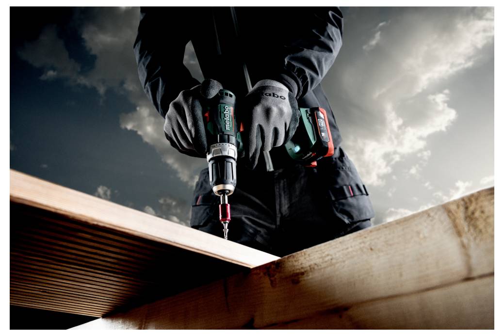 Eine Person, die mit einer kabellosen Bohrmaschine eine Holzplanke unter einem bewölkten Himmel befestigt, trägt Handschuhe und eine Jacke, was Bauarbeiten veranschaulicht.