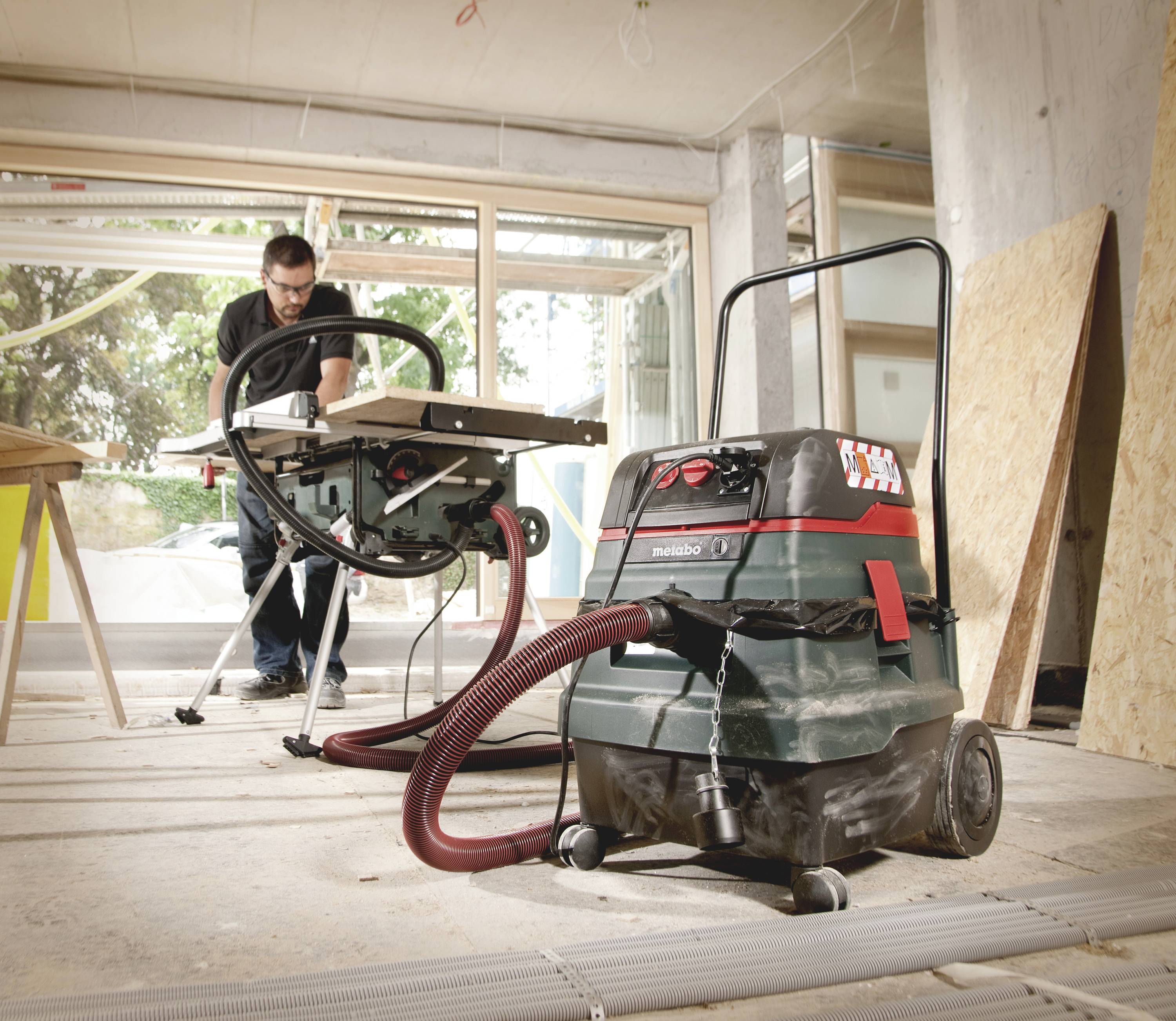 Ein Mann bedient eine Kreissäge in einer Werkstatt, wobei ein Absaugsystem zur Staubsammlung angeschlossen ist. Holzplatten und Baumaterialien sind zu sehen.