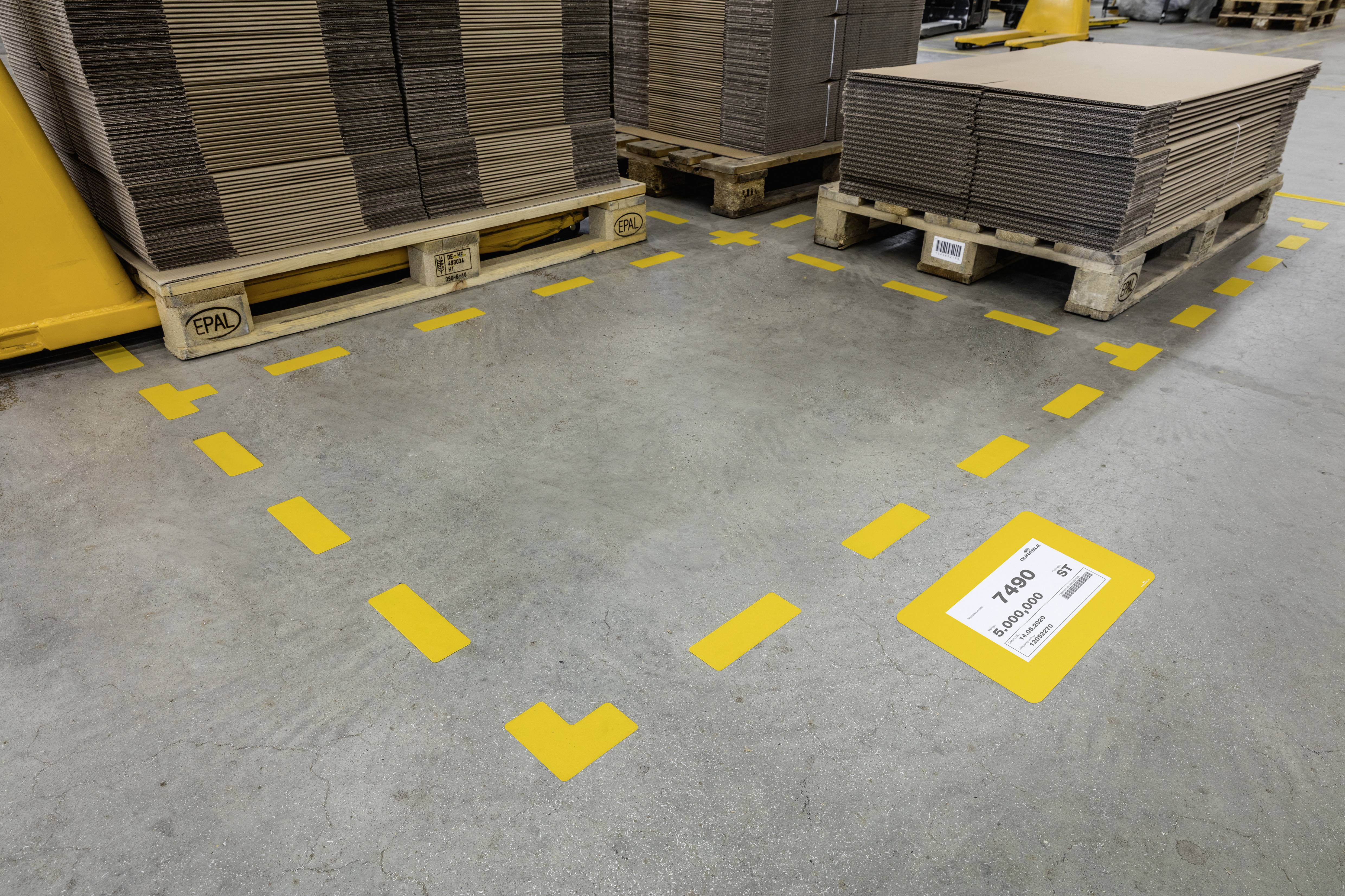 Mehrere Pappkartons auf Paletten, sicher in einem markierten gelben Bereich auf einem Lagerboden platziert. Ein Schild weist auf Sicherheitsabstand hin.