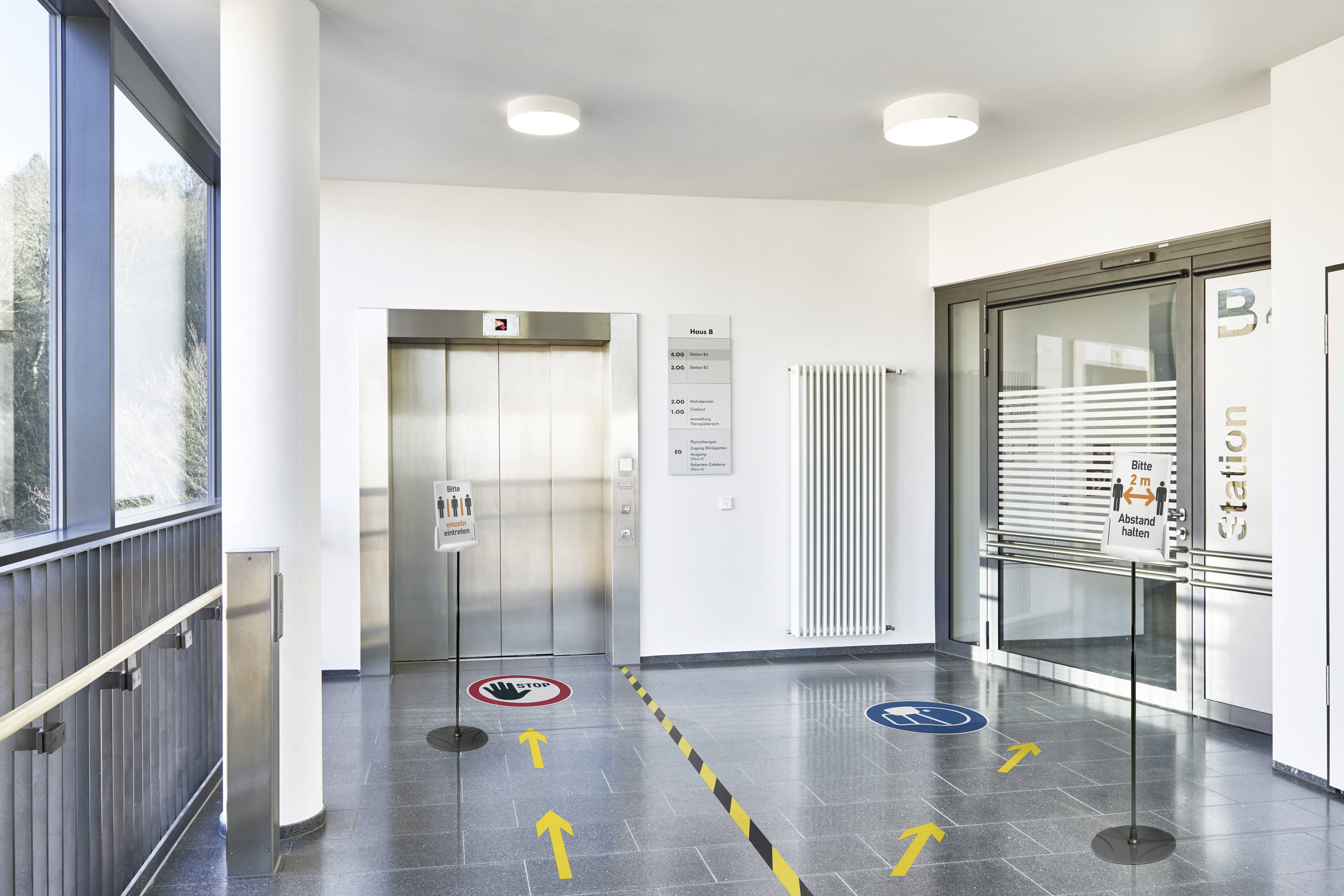 Eingangsbereich mit Aufzug. Bodenmarkierungen zeigen Richtungen an. Hinweisschilder für Hygiene und Barrierefreiheit sind sichtbar.