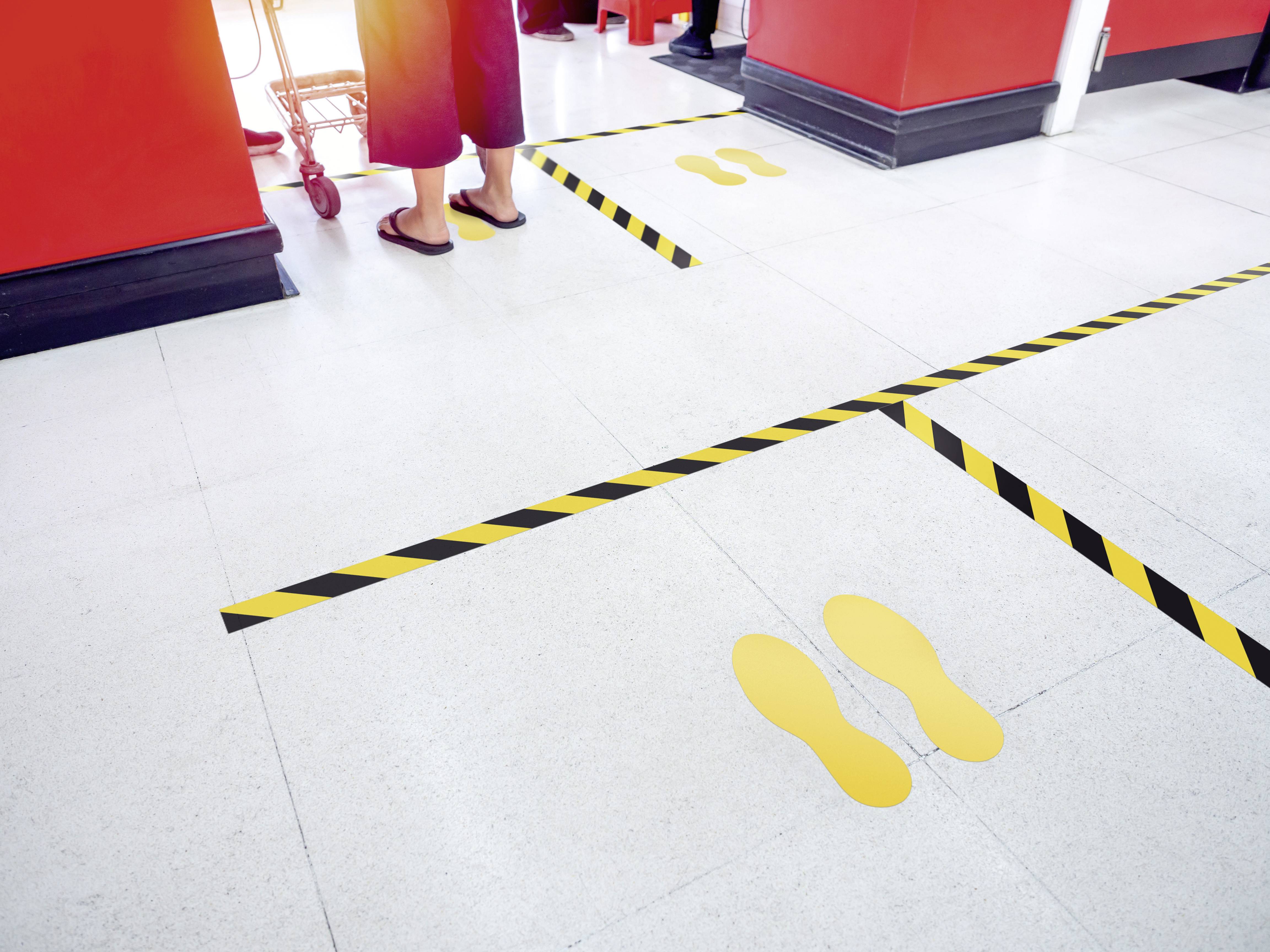 Gelbe Fußabdrücke kennzeichnen den Wartebereich in einem Geschäft vor der Kasse, getrennt durch gelb-schwarze Abstandsmarkierungen.