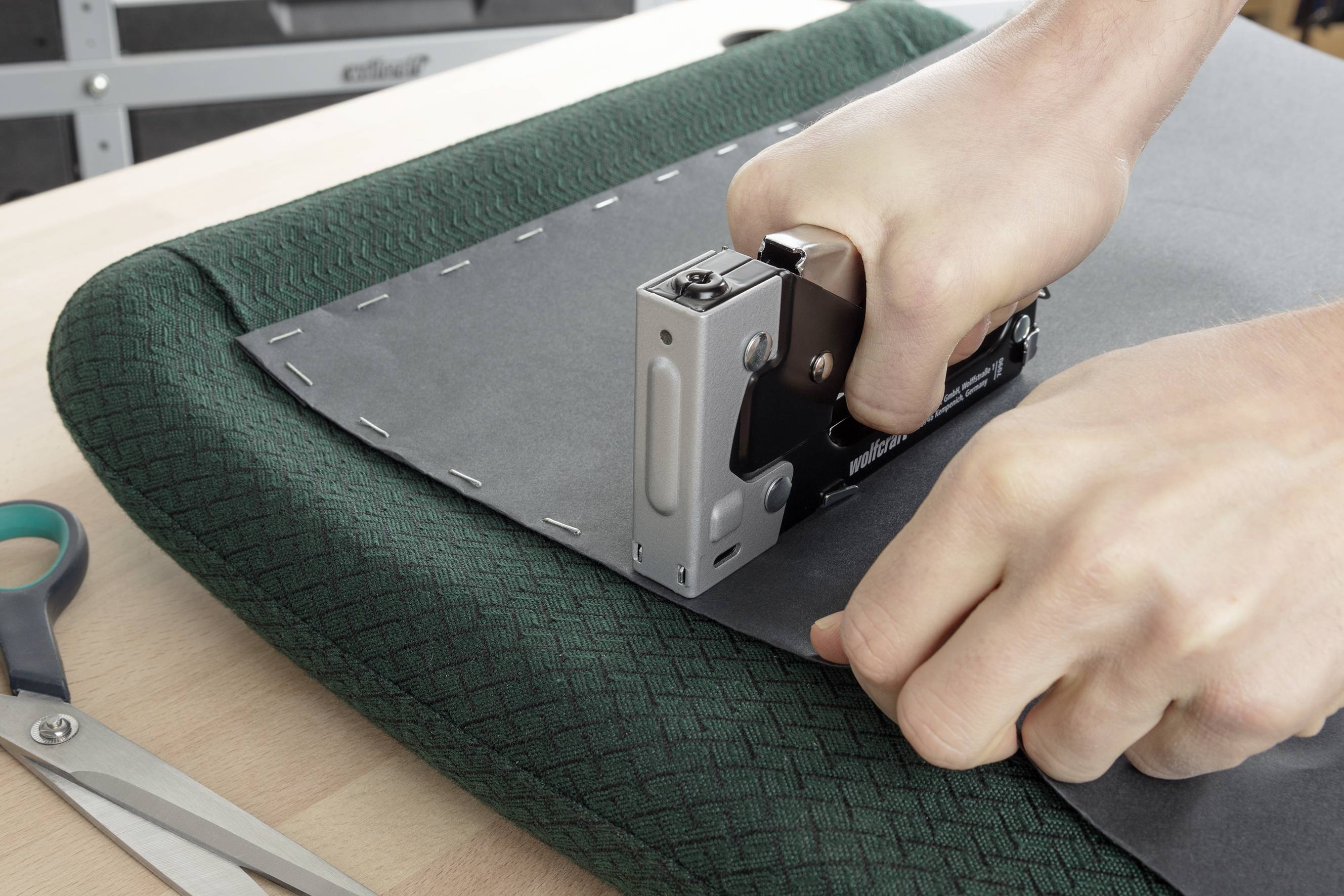 Eine Person befestigt mit einem Tacker einen grauen Stoffbezug auf einem Polster. Eine Schere liegt daneben auf dem Tisch.