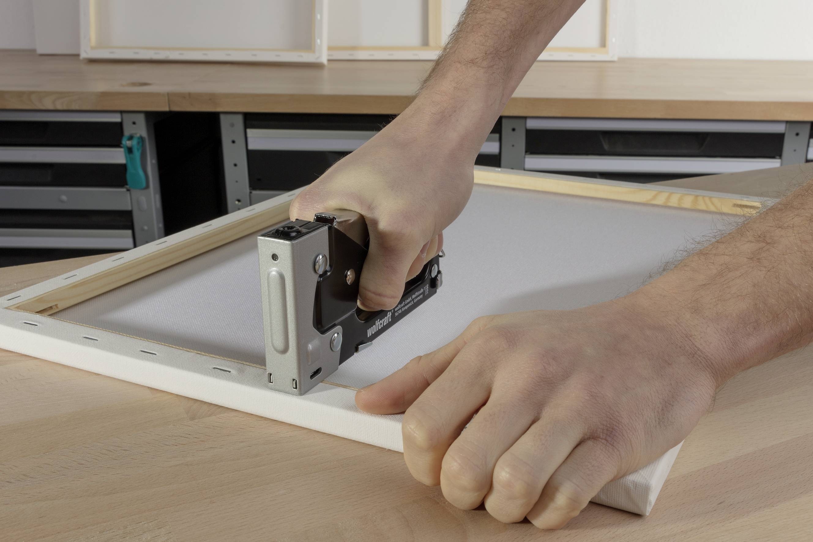 Zwei Hände befestigen eine Leinwand mit einem Tacker auf einem Holzrahmen. Der Tisch im Hintergrund ist aus Holz.