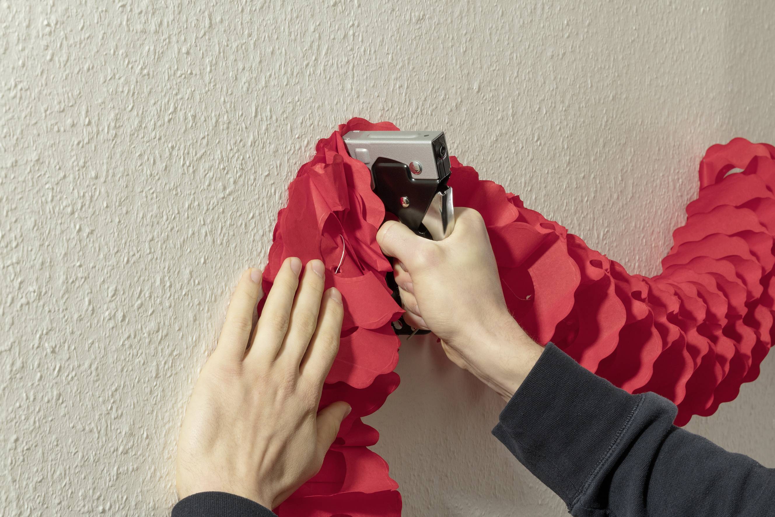 Person befestigt rote Dekoration mit Tacker an der Wand. Hände halten Dekoration fest.