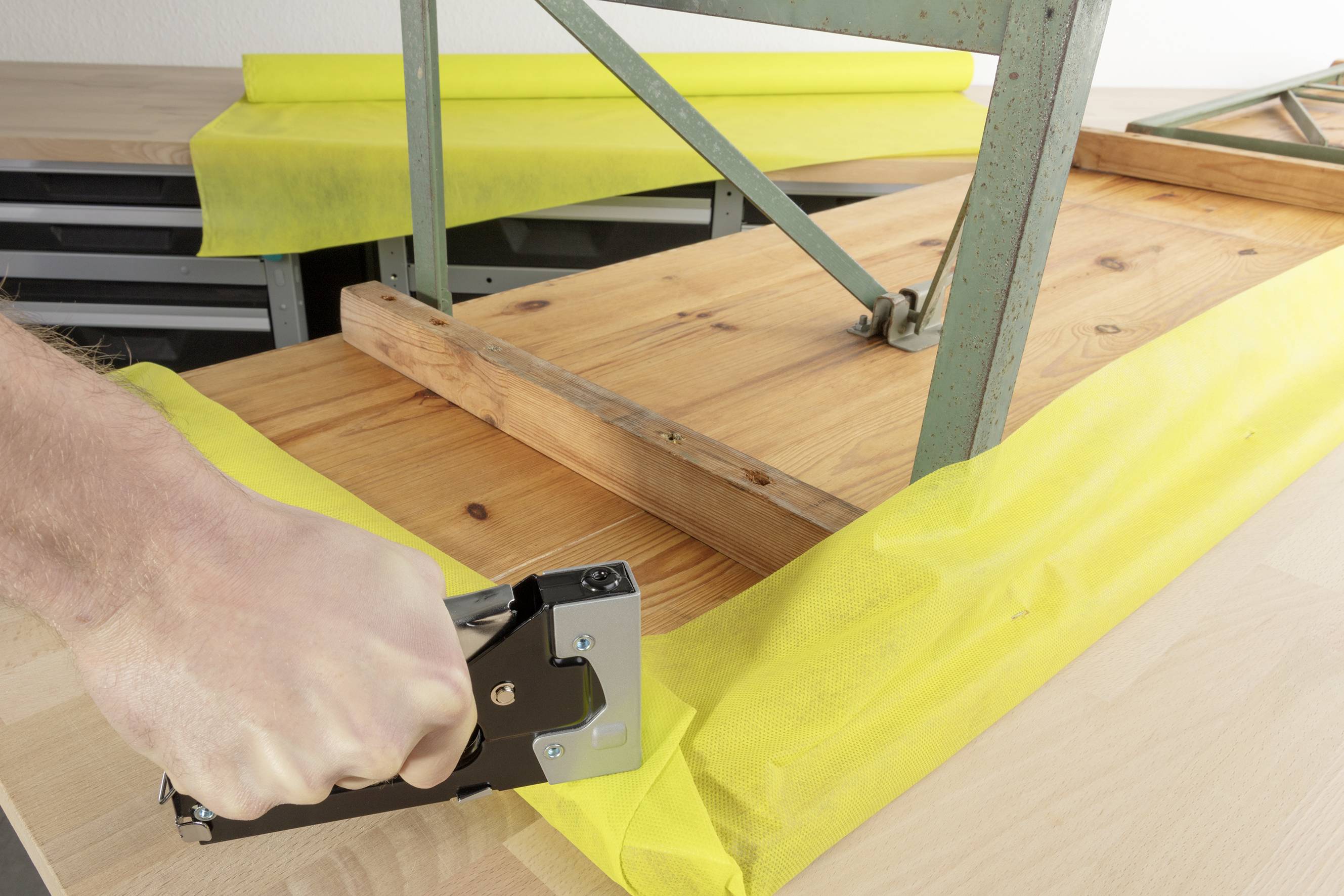 Eine Hand verwendet einen Tacker, um gelben Stoff an einen Holztisch zu befestigen. Ein Metallrahmen steht auf dem Tisch.