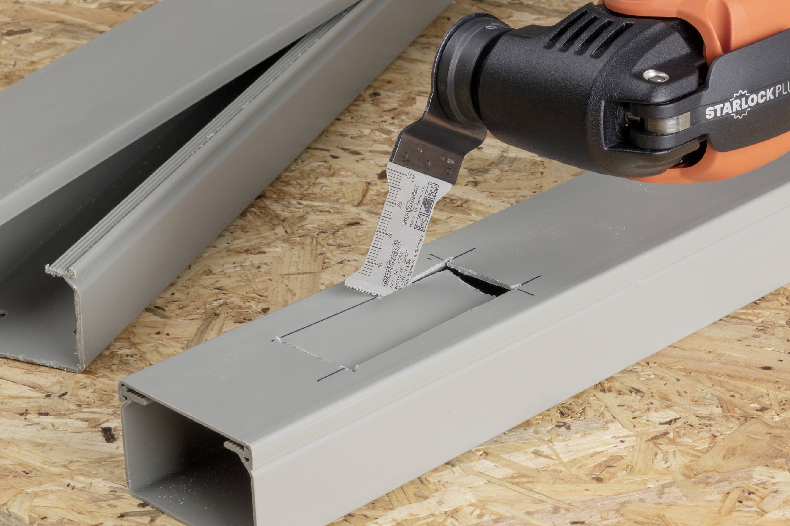 Ein oszillierendes Multitool schneidet präzise in ein graues Kunststoffrohr, das auf einer Holzplatte liegt.