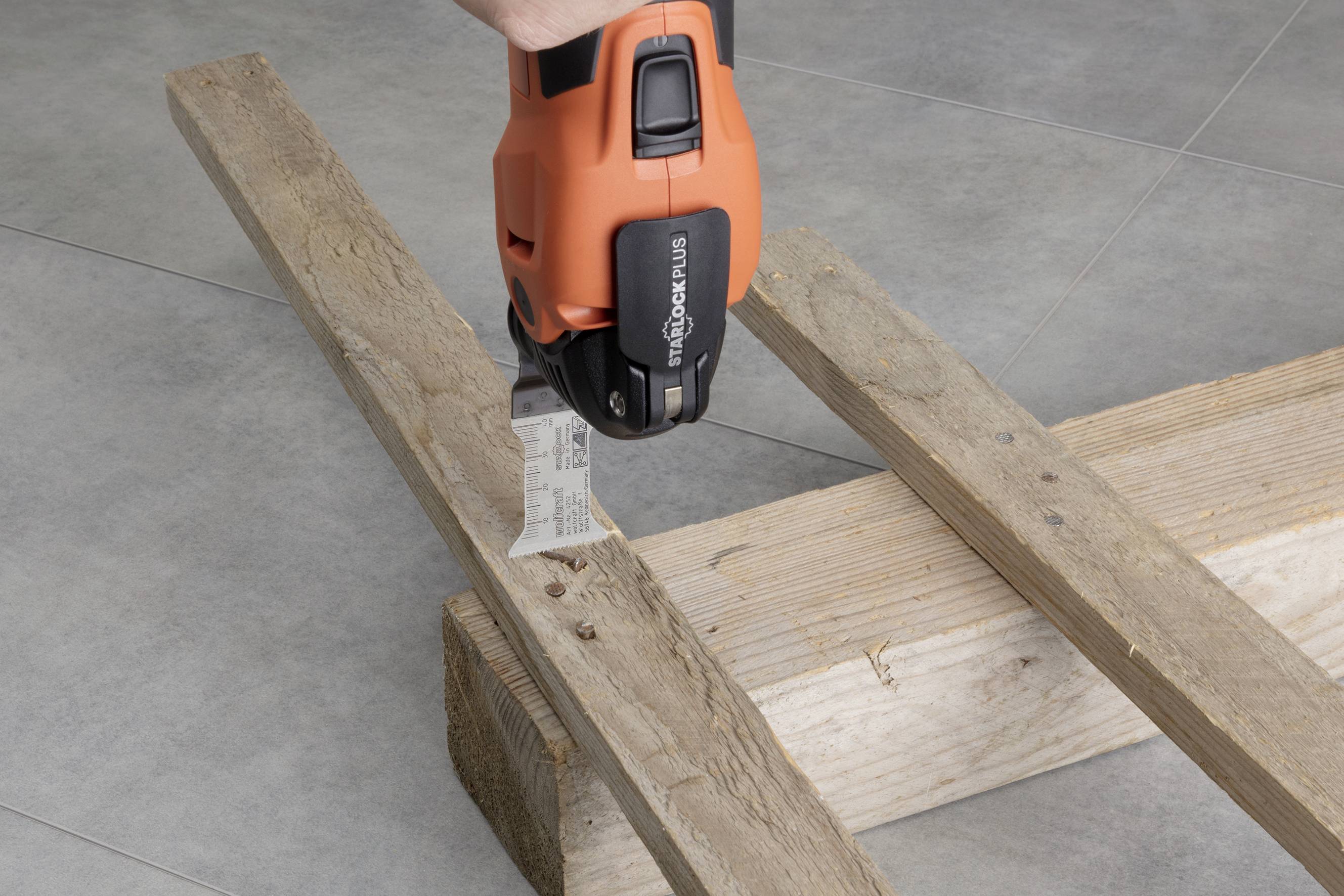 Eine orangefarbene Elektrosäge durchtrennt ein Stück Holz, das auf zwei weiteren Holzlatten liegt.