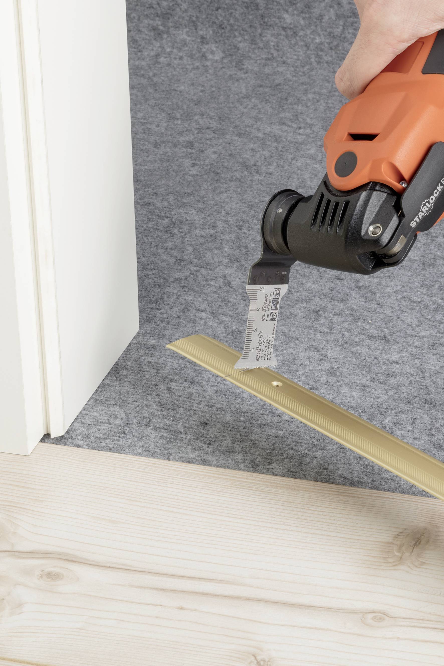 Eine Person schneidet mit einer oszillierenden Säge eine goldene Metallschiene auf einem Holzfußboden neben einem Türrahmen.