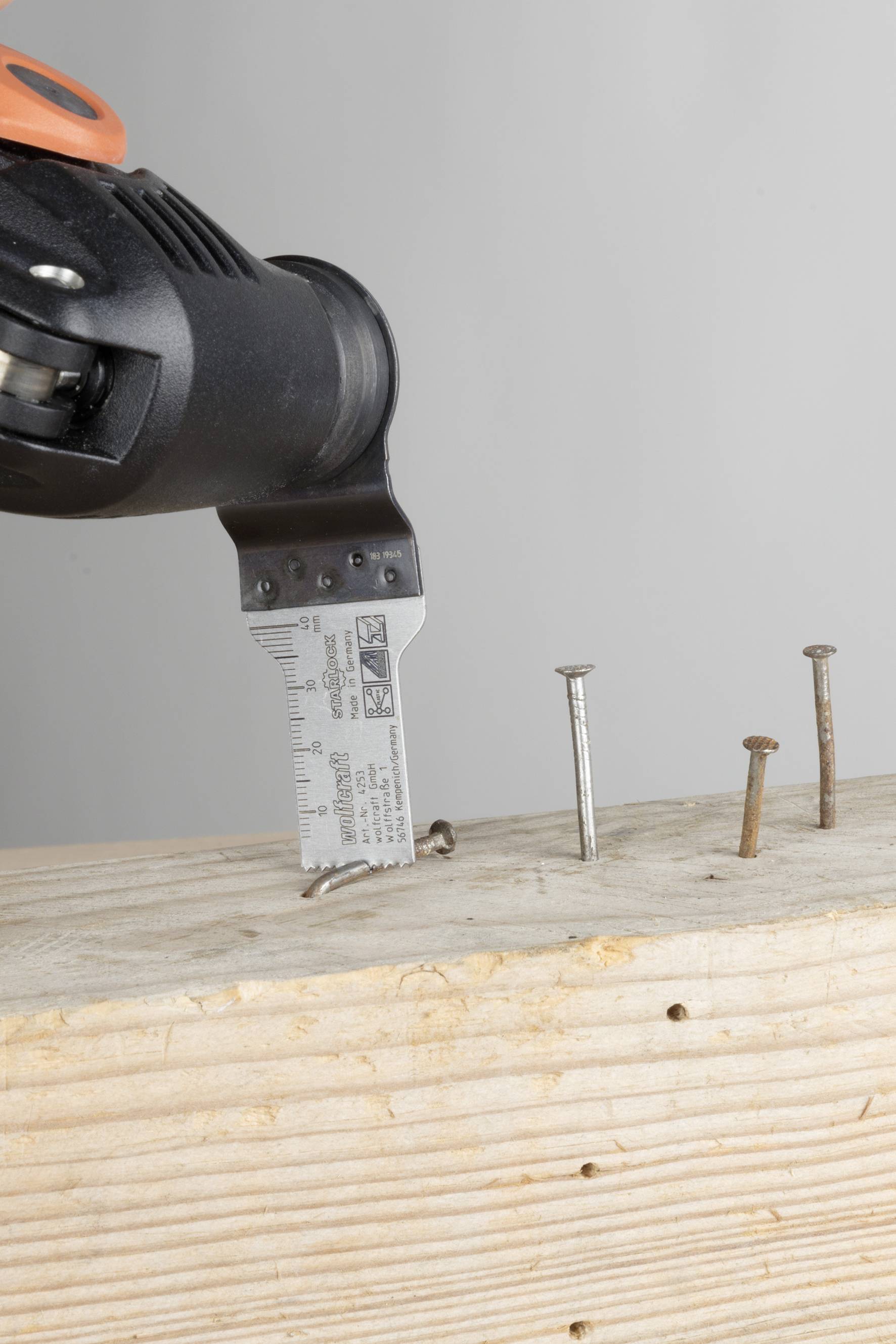 Ein elektrischer Schraubendreher bewegt eine gebogene Schraube in ein Holzbrett, neben mehreren versenkten Schrauben.