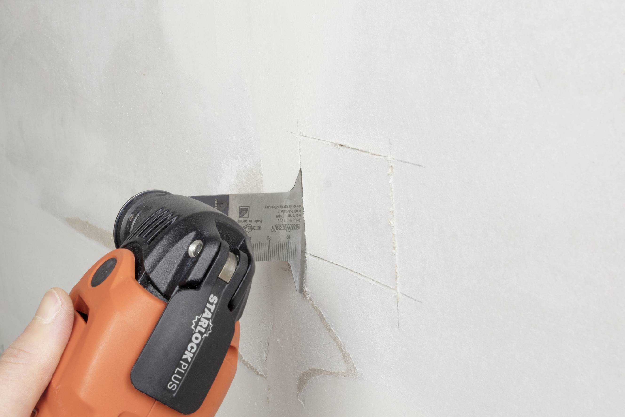 Eine Person schneidet mit einem orangenen Elektrowerkzeug ein quadratisches Loch in eine Trockenbauwand.