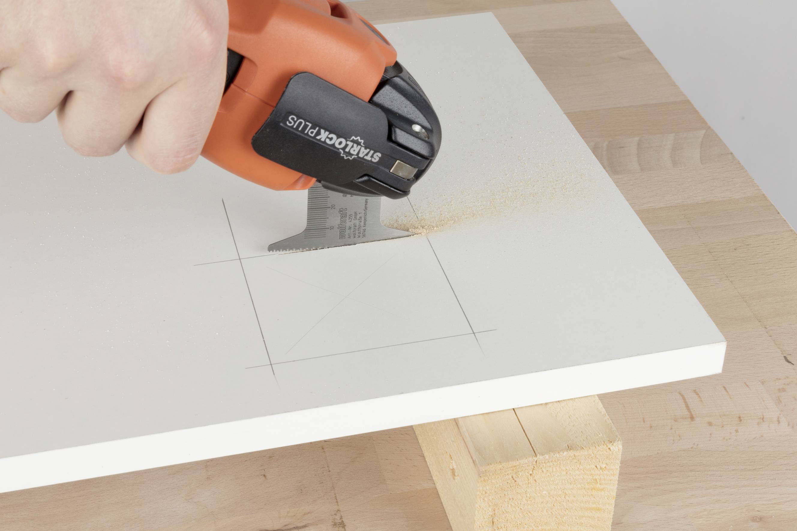 Hand hält oszillierendes Multifunktionswerkzeug, sägt rechteckigen Ausschnitt in weißem Holzbrett auf Werkbank.