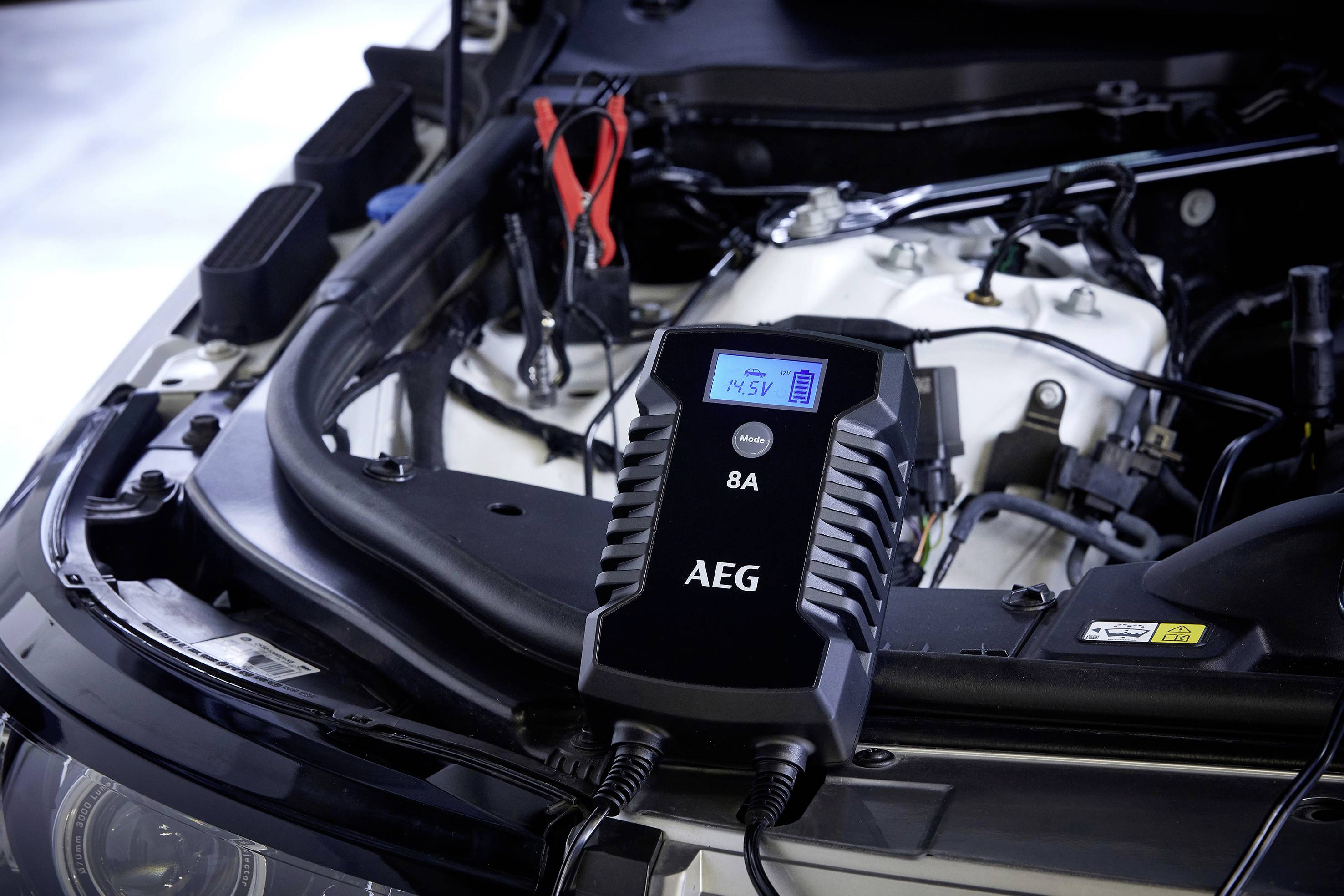 Batterieladegerät der Marke AEG, angeschlossen unter der geöffneten Motorhaube eines Autos. Ladestatus wird auf einem Display angezeigt.